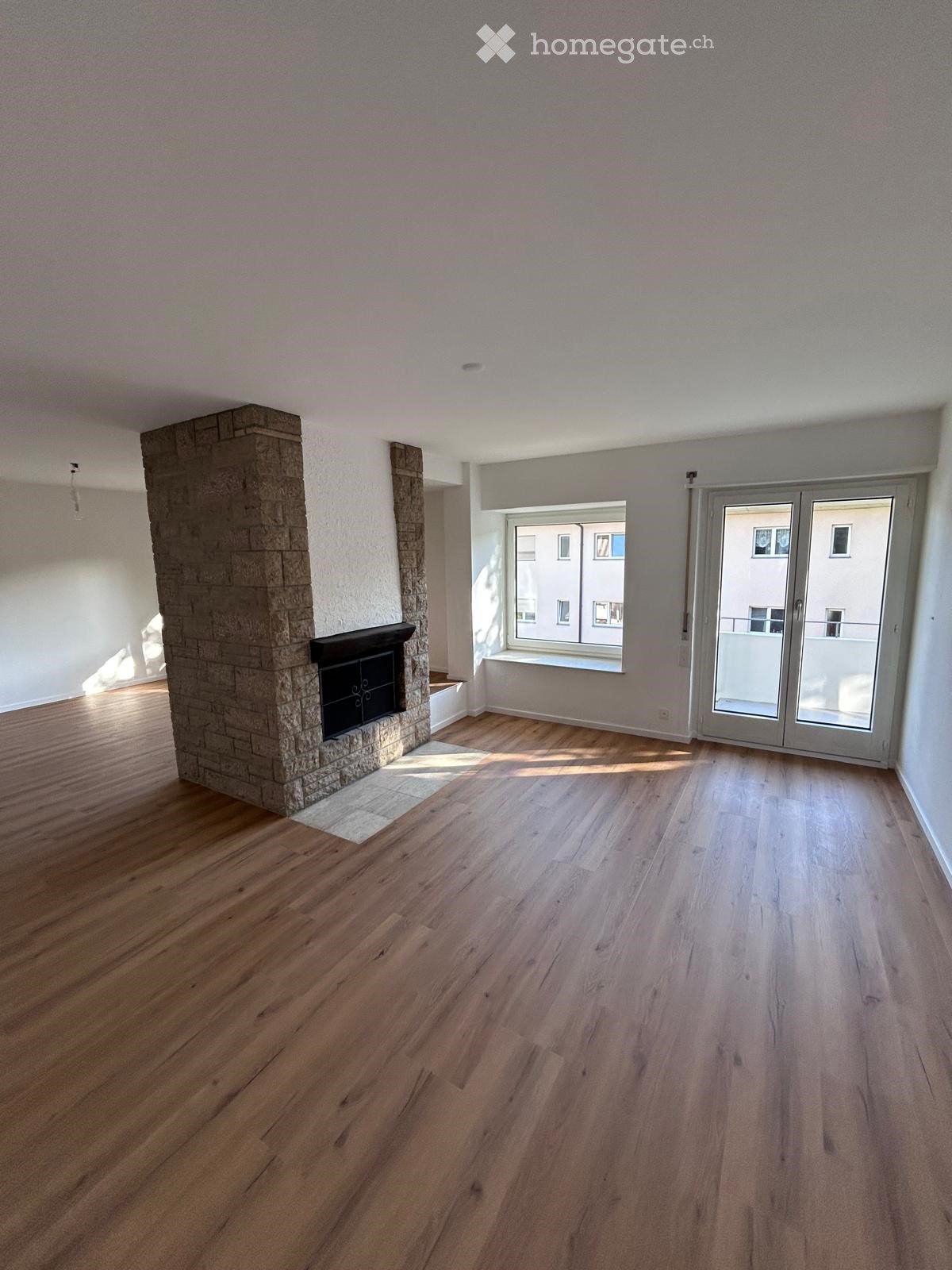 Empty room with wooden flooring, a stone fireplace, large window, and sliding glass door leading to a balcony.