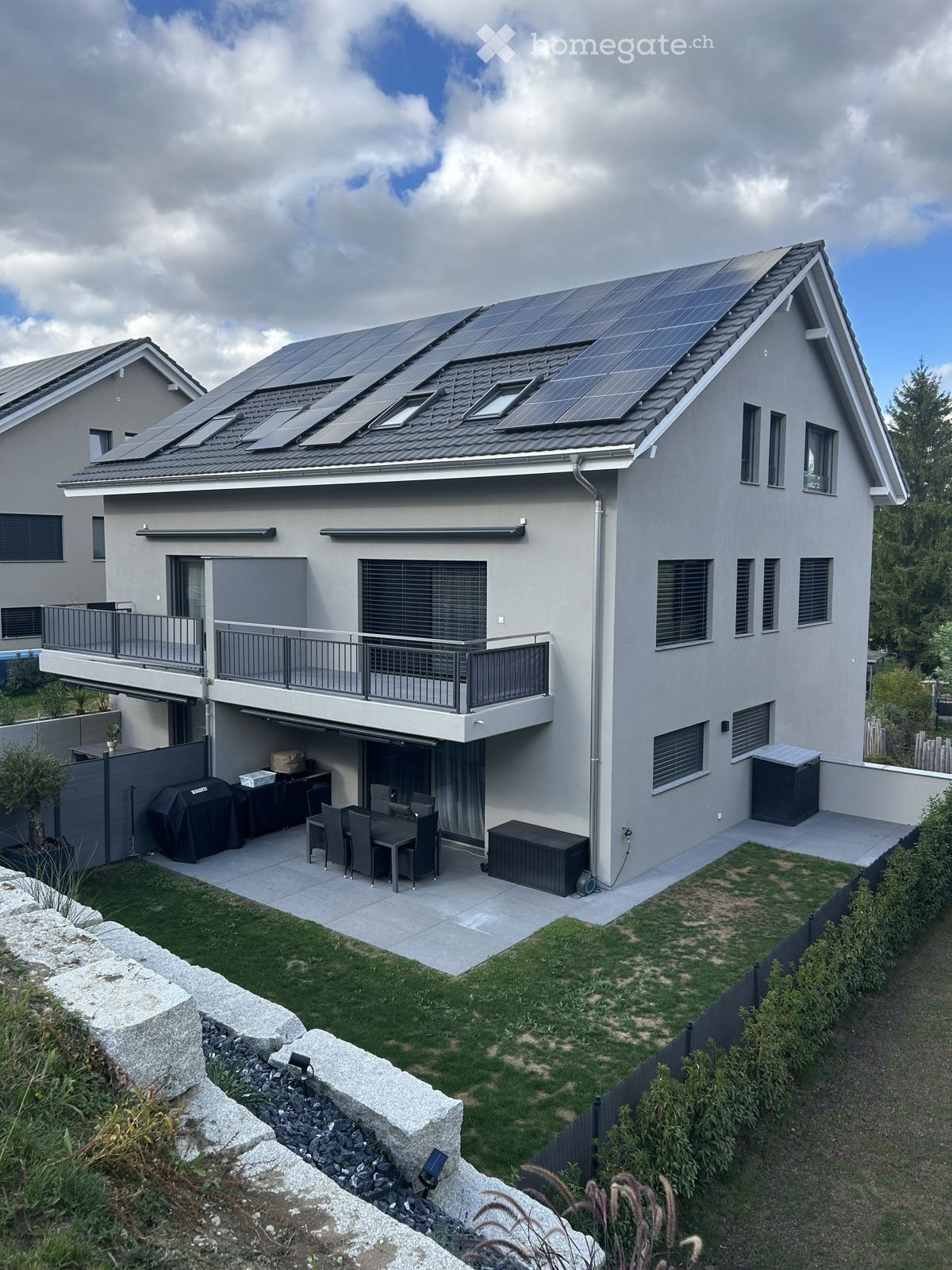 Modern two-story house with solar panels on the roof, large windows, balcony, garage, and outdoor patio with dining set and grill.