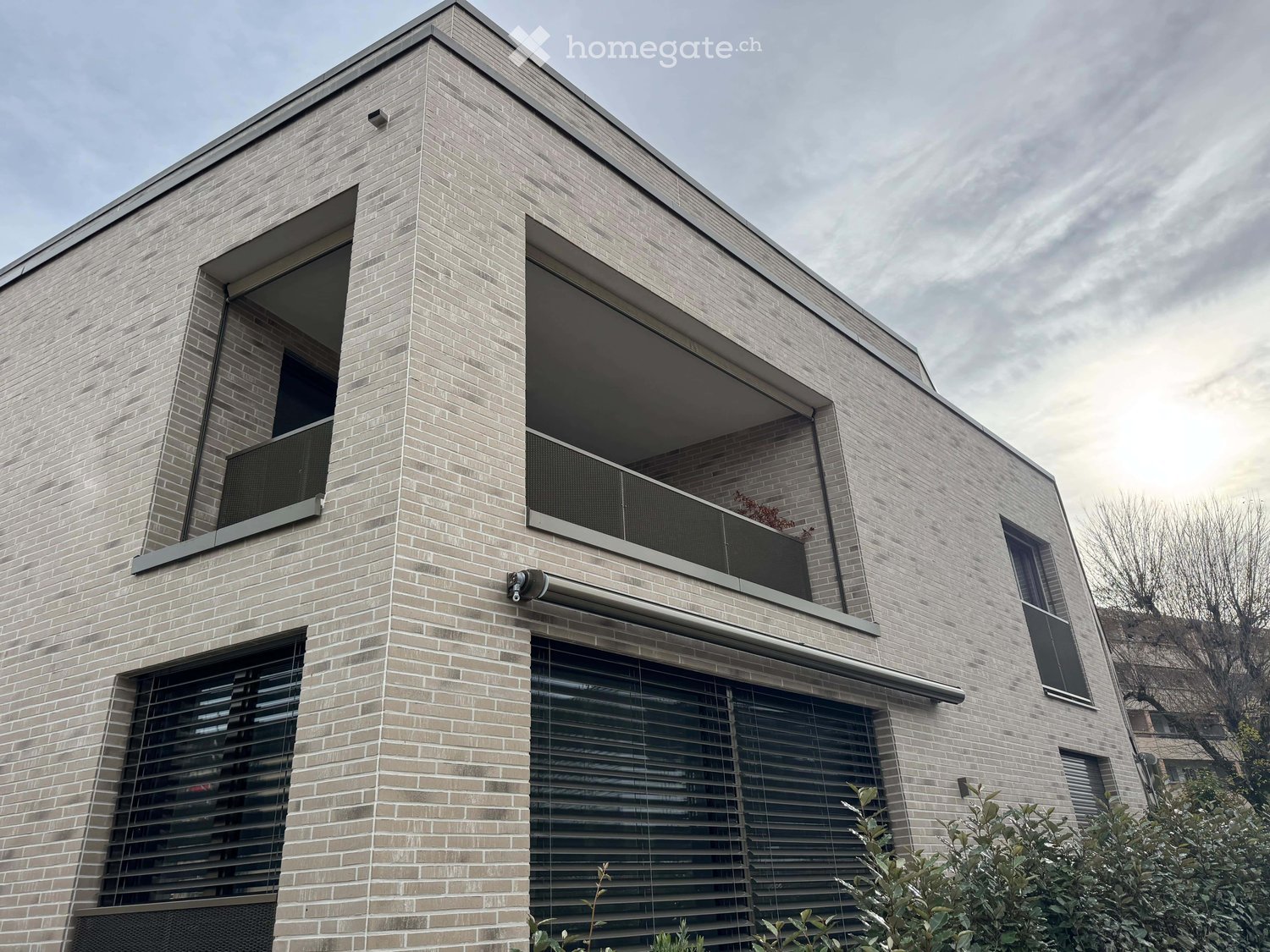 Modern two-story house with light gray brick facade, large windows with blinds, and a balcony on the upper level.