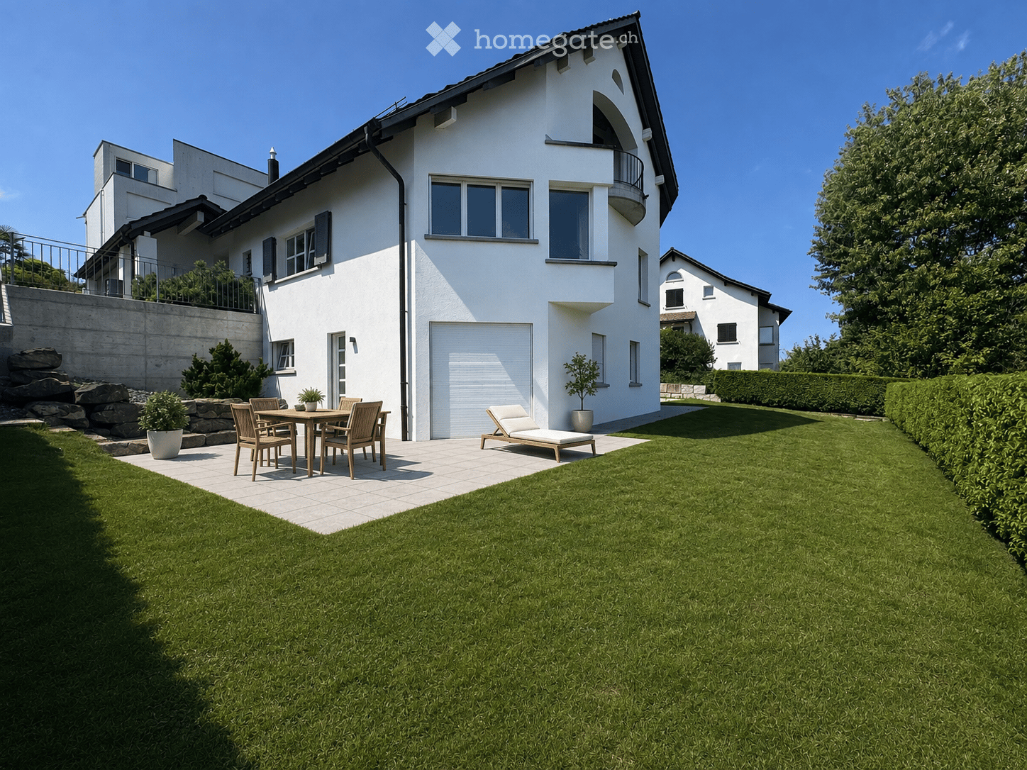 Well-maintained garden with a modern white house in the background, featuring a patio area with a wooden table, chairs, a sun lounger, and potted plants.