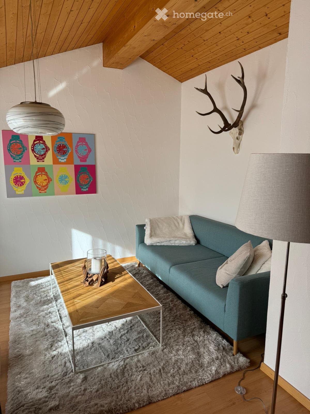 Living room with wooden ceiling, white walls, blue couch, wooden coffee table, white rug, lamp, deer antlers mounted on wall, colorful watch art on wall