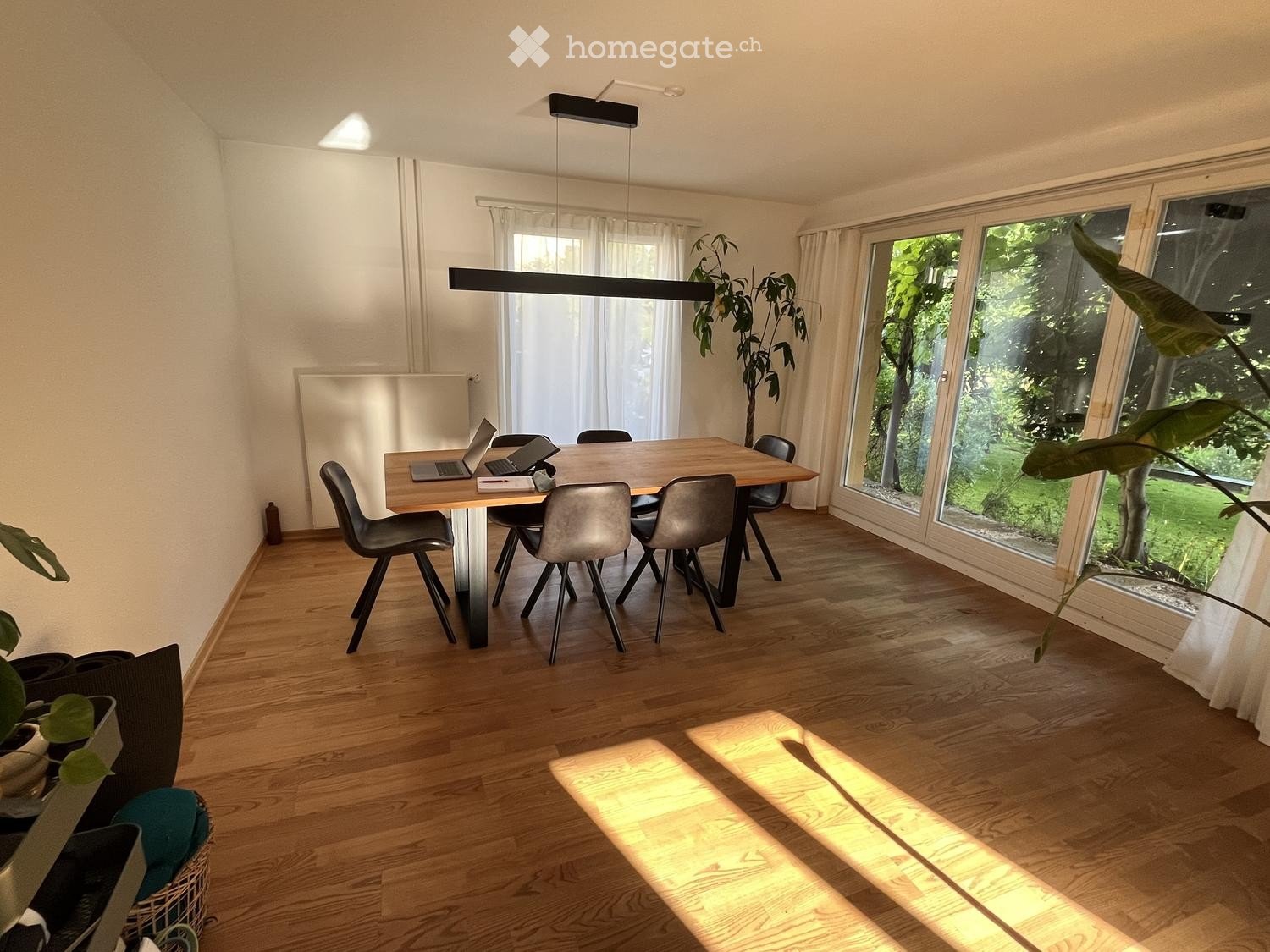 Bright dining area with wooden table, six chairs, laptops, and large windows overlooking greenery.