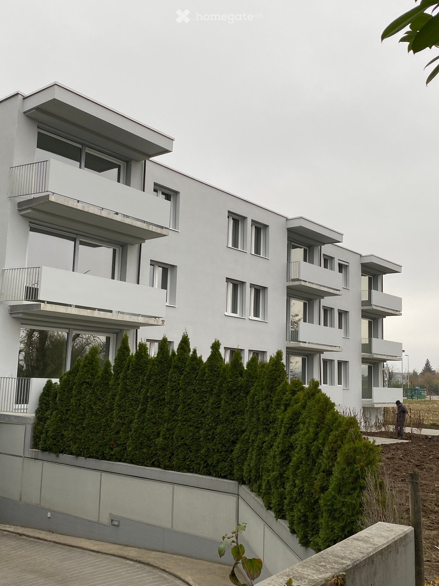 Modern white multi-story building with multiple balconies and large windows, surrounded by neatly trimmed evergreen shrubs and a raised ground floor entrance.