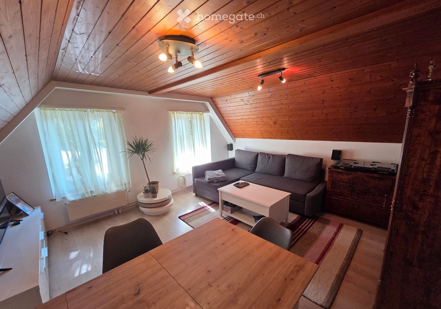 A cozy living room with wooden beams on the ceiling, a grey sofa, a white coffee table, a wooden dining table, and a potted plant near the window.