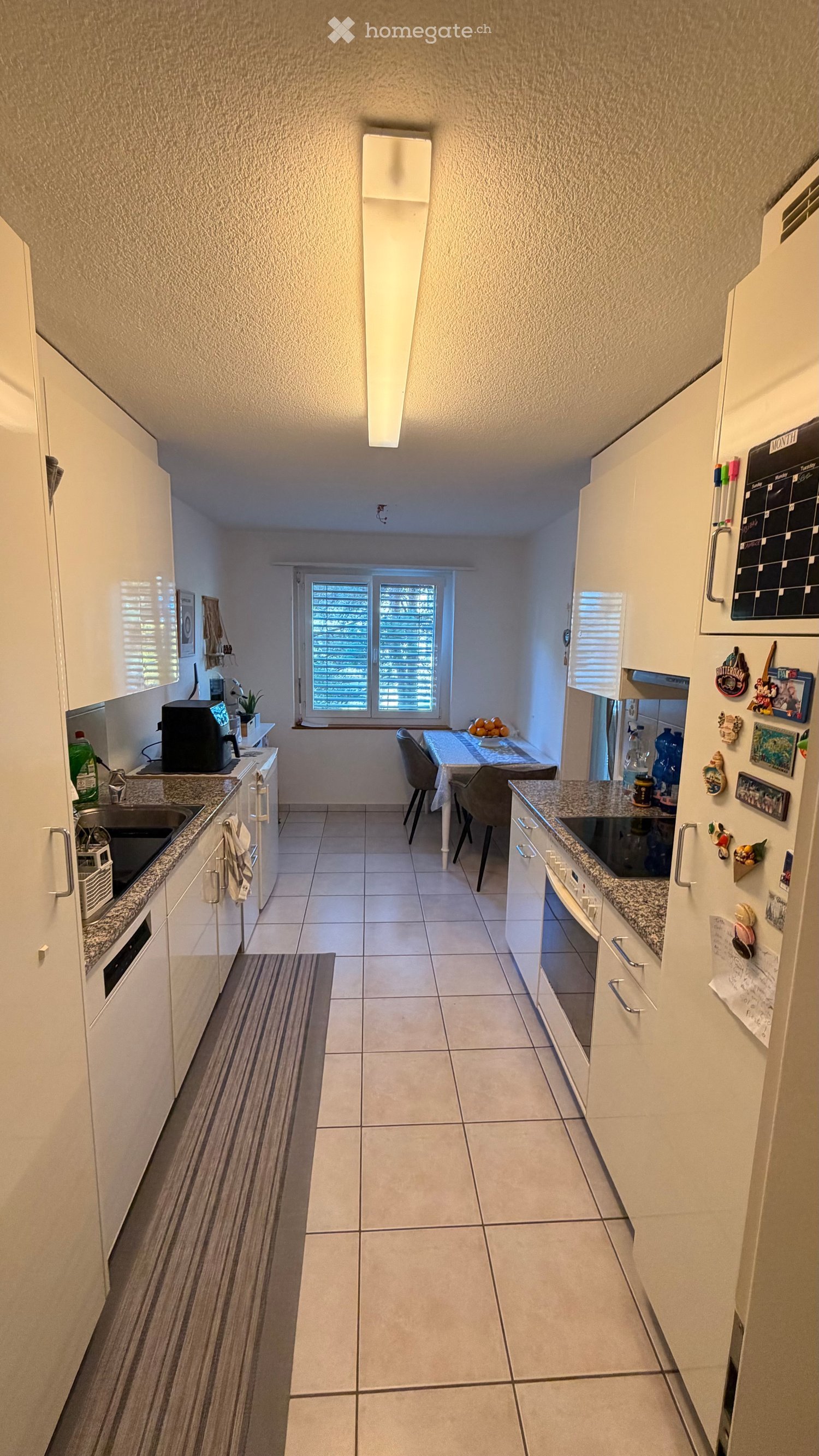 Modern kitchen with white cabinets, granite countertops, and a dining area with a table and chairs. Features include a refrigerator with magnets, a coffee maker, and a calendar on the wall.