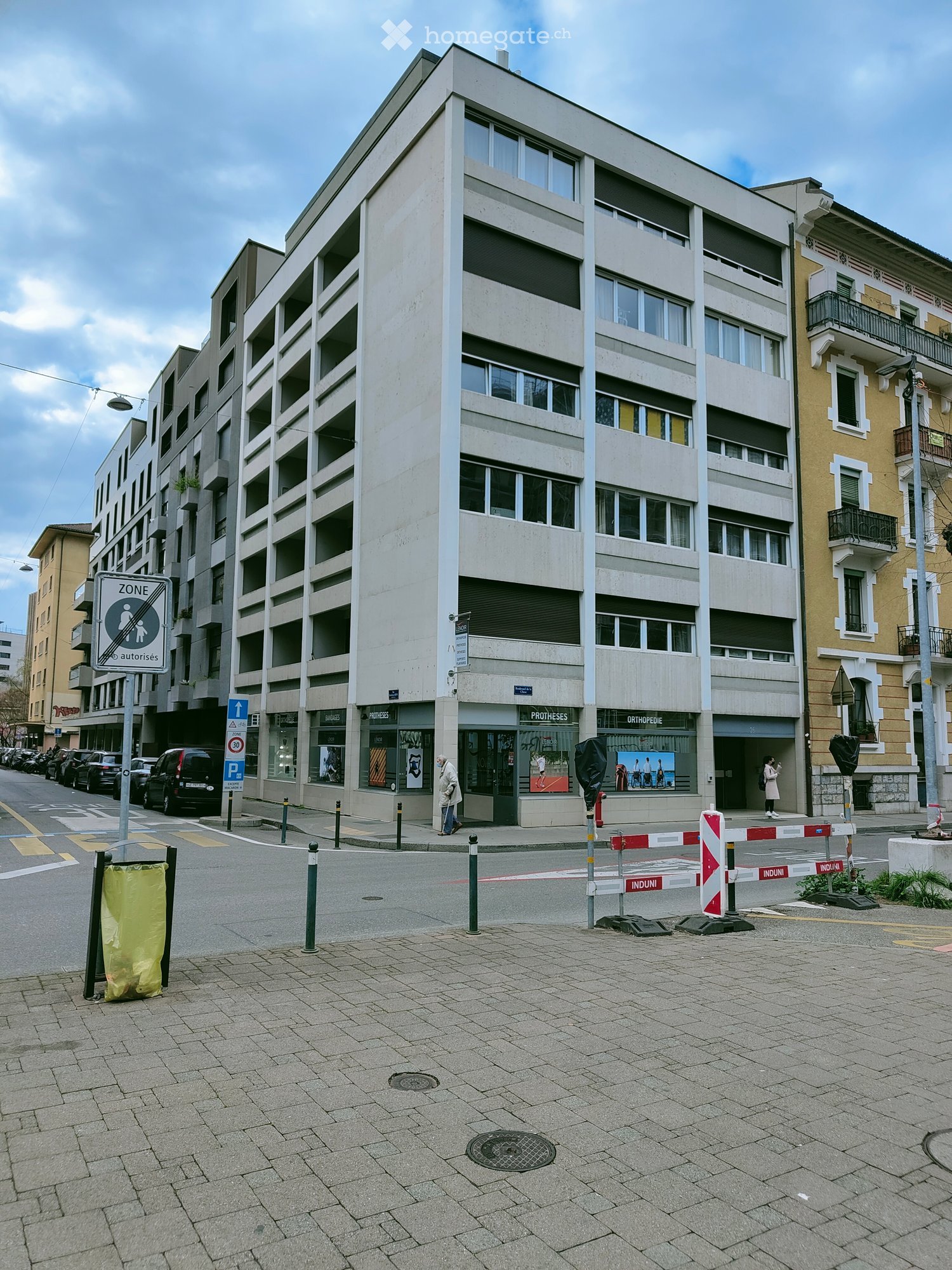 A modern multi-story building with large windows and commercial storefronts at the ground level, located on a paved street with traffic signs and barriers.