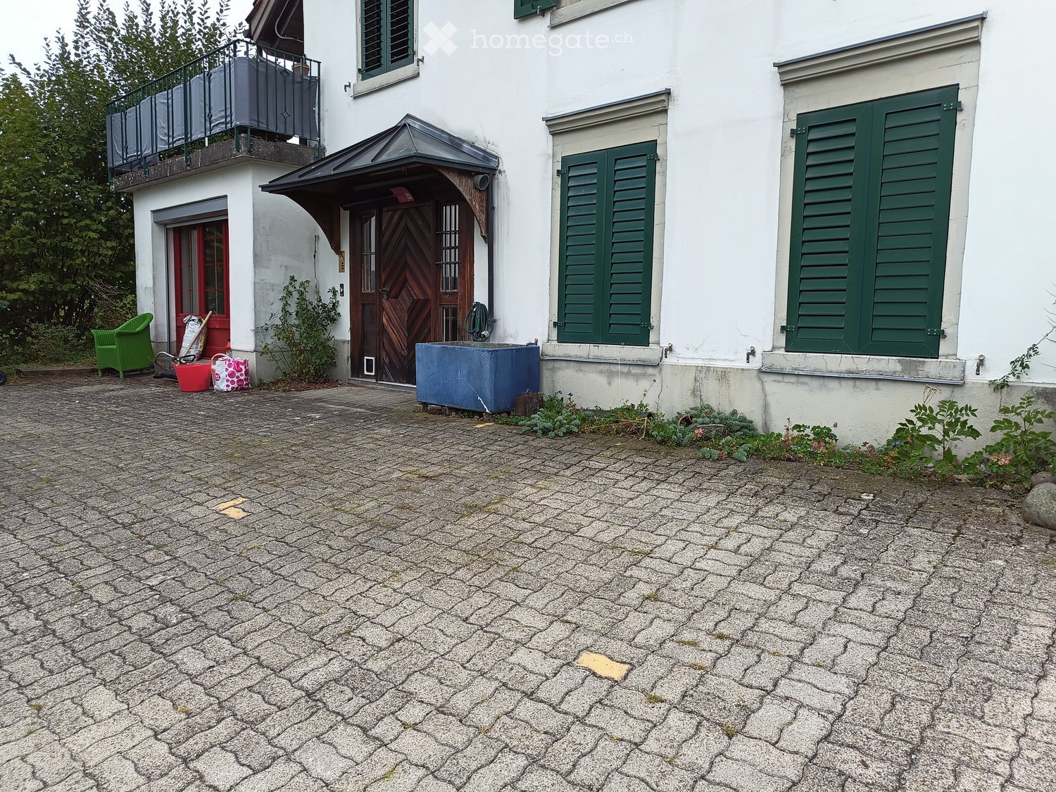 Two-story white building with green shutters, a balcony, and a wooden entrance door. The ground is paved with cobblestones, and there are some outdoor items near the entrance.