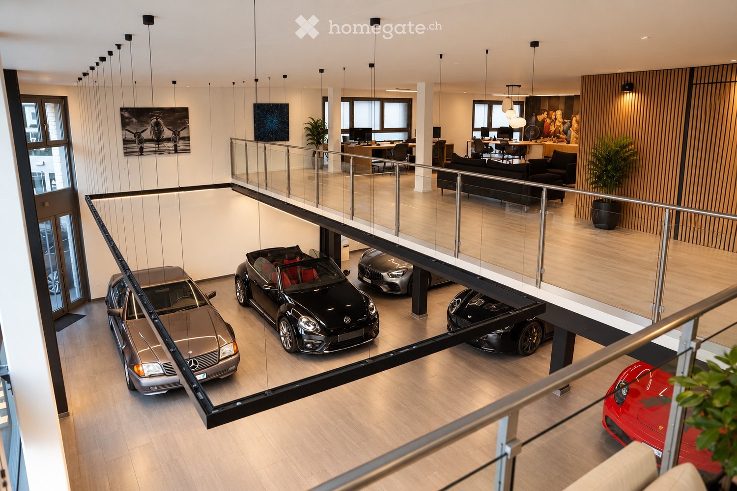 Modern, multi-level showroom with a sleek design, featuring a glass balcony overlooking a lower level with several luxury cars displayed. The upper level includes a seating area with a sofa, chairs, a table, and a large screen, along with office desks and