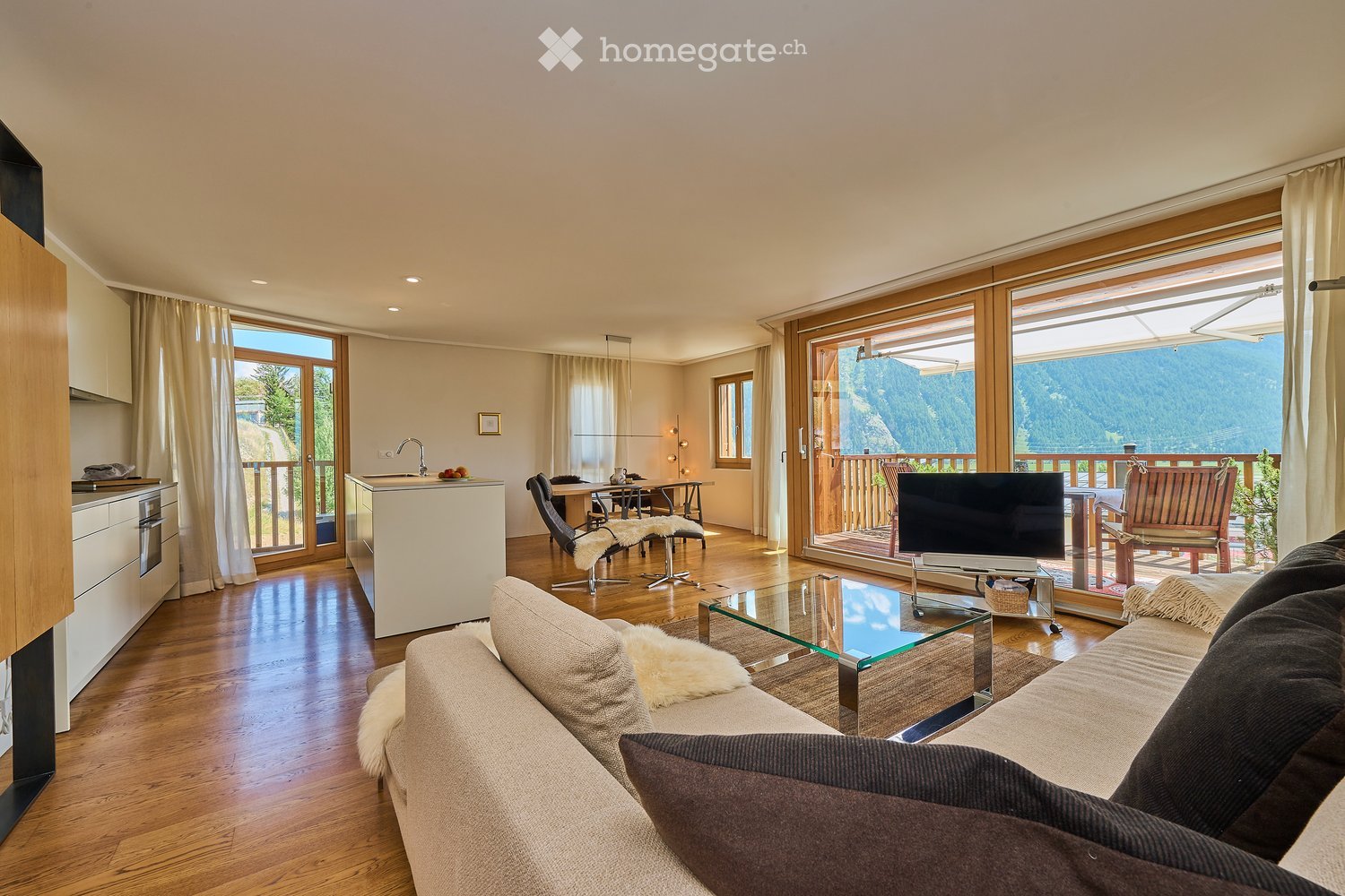 Open-plan living and dining area with a modern sofa, coffee table, TV, and a kitchen island with a sink and faucet. Large windows offer a scenic mountain view.