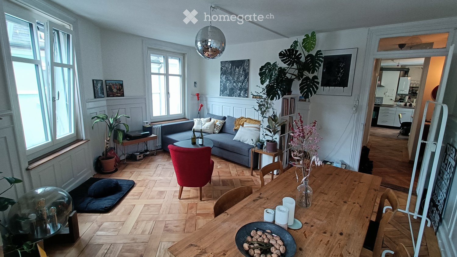 Bright living room with wooden flooring, large windows, a grey sofa with pillows, a coffee table, a red chair, a dining table with chairs, and decorative plants.
