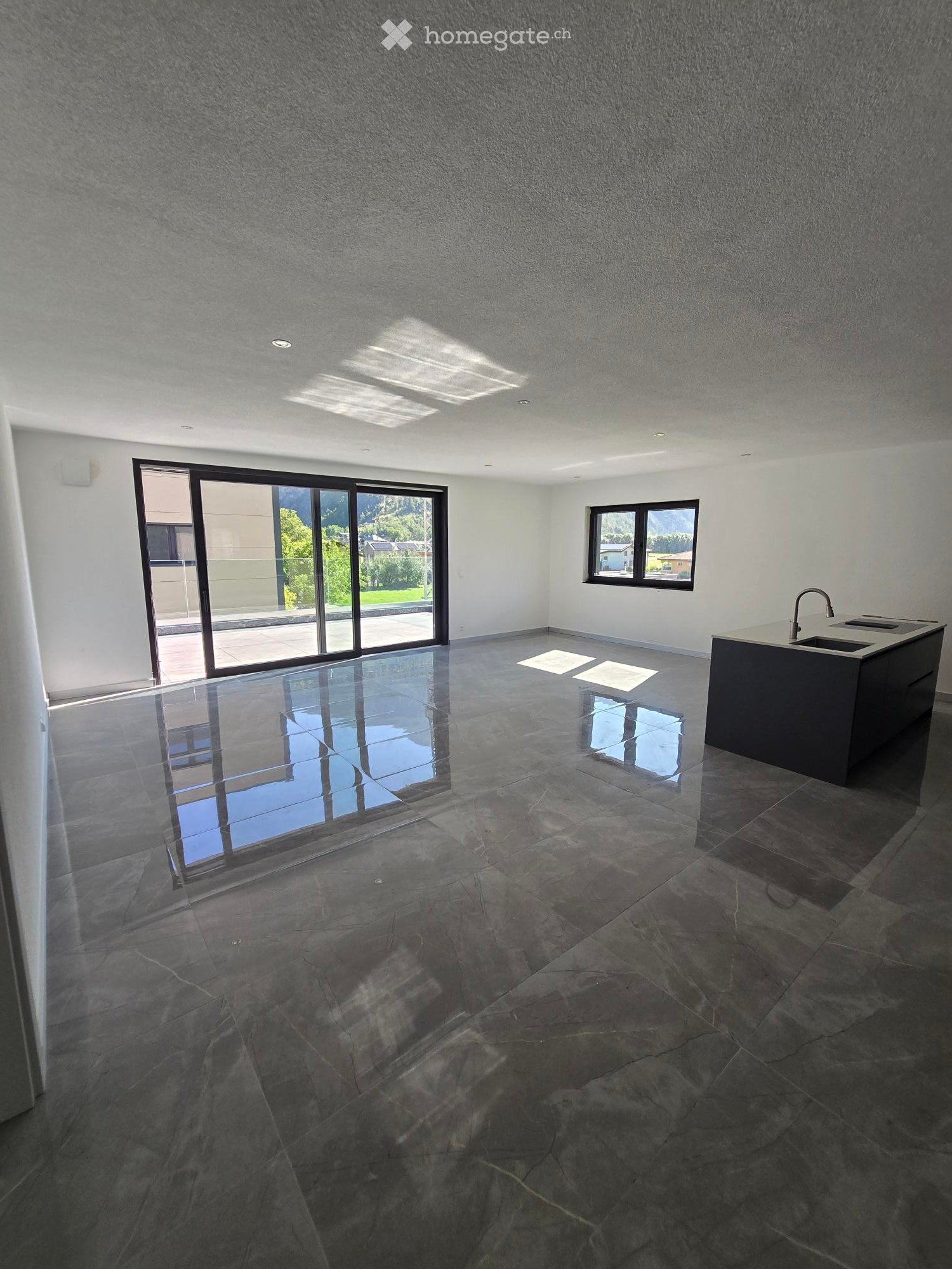 Empty room with dark stone tiles, large windows, and a modern kitchen island with a sink.