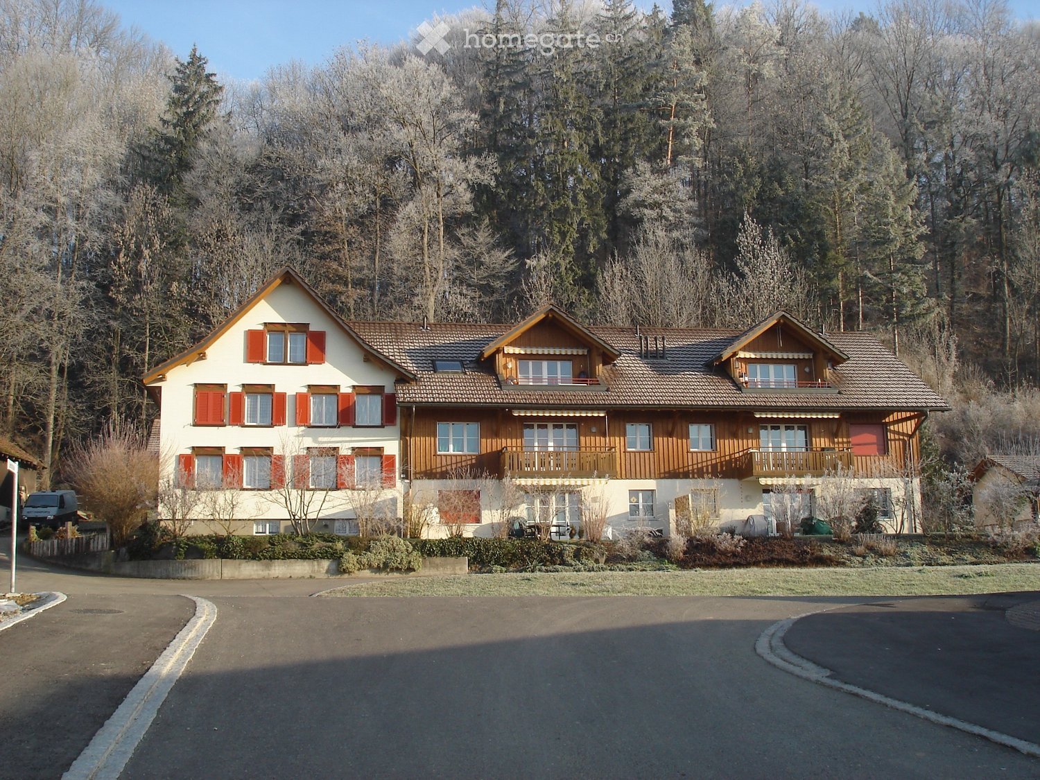 Two-story house with red shutters, wooden balconies, and a tiled roof, surrounded by trees and a driveway.