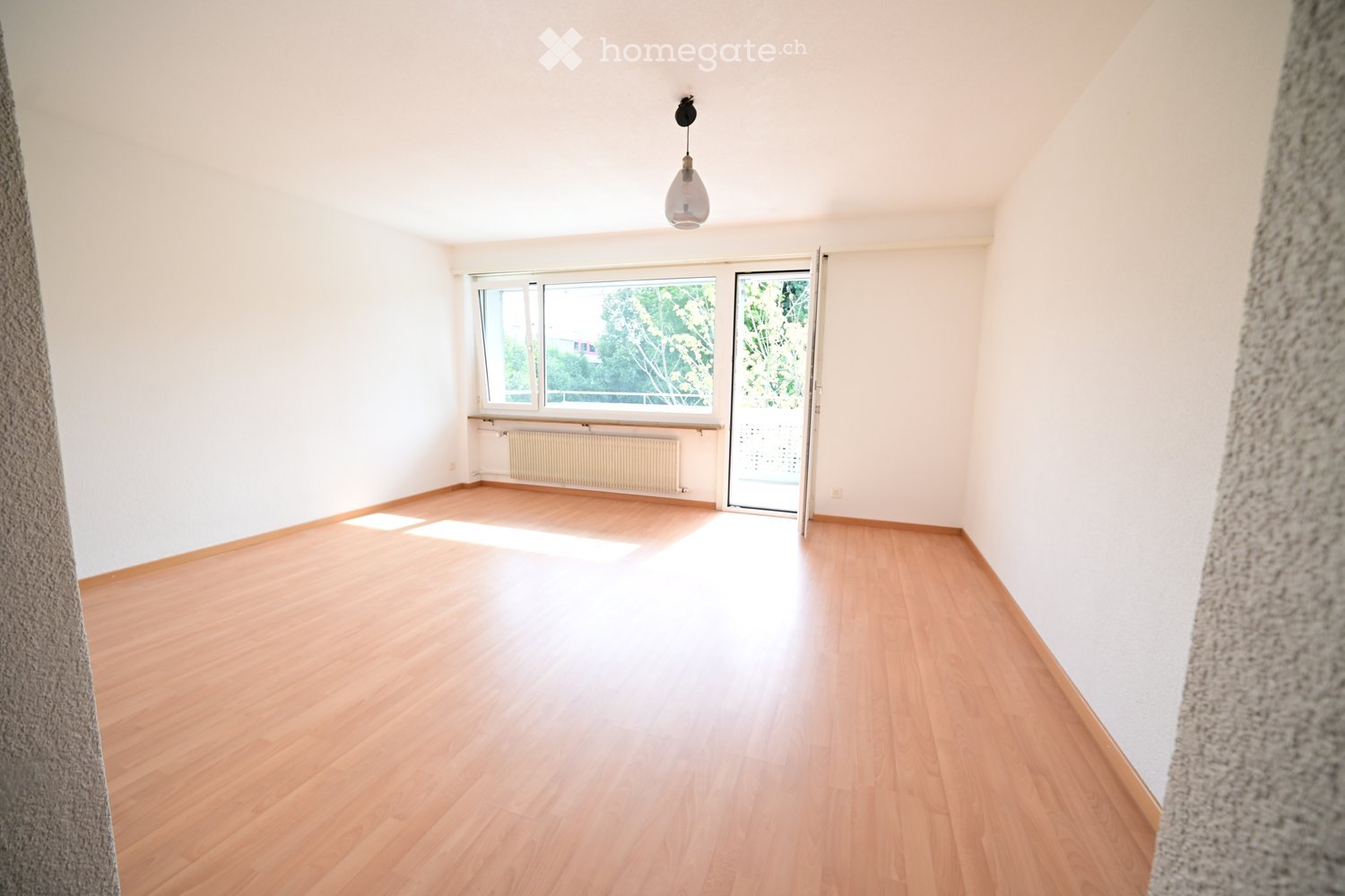 Empty room with wooden flooring, white walls, large windows, and a sliding glass door leading to a balcony.