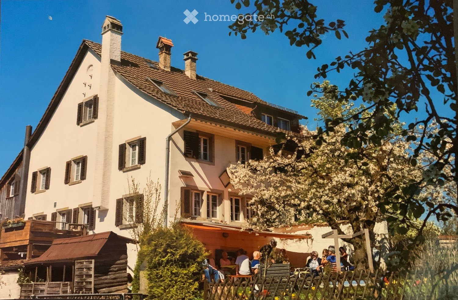 A two-story house with a tiled roof, multiple chimneys, and several windows with shutters. The house has a balcony and is surrounded by greenery, including a blooming tree and a wooden fence. There is an outdoor seating area with tables and chairs where p