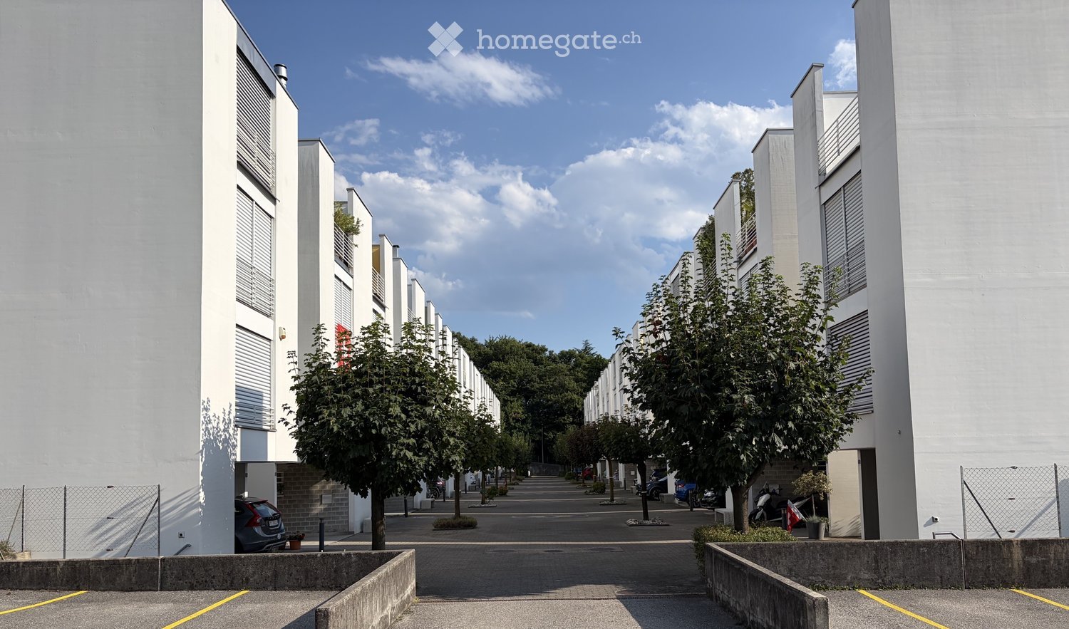 Modern residential building complex with white facades, multiple floors, and a central walkway lined with trees and parked cars.