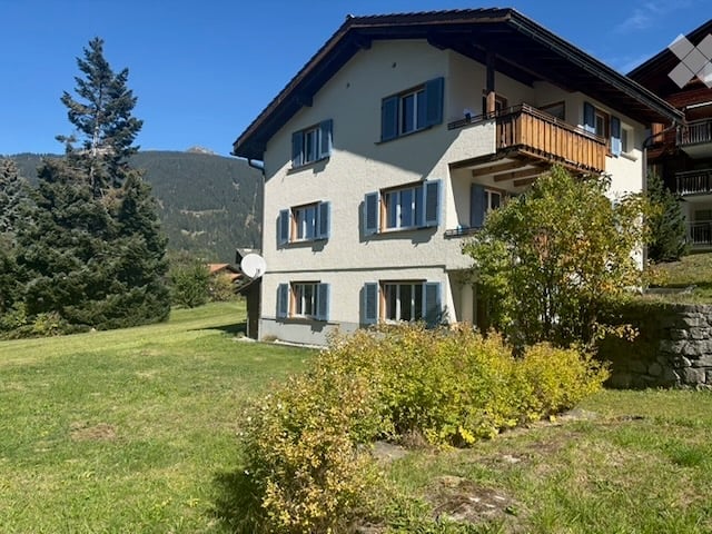 Two-story house with white walls, blue shutters, and a balcony overlooking a green lawn and mountain view.