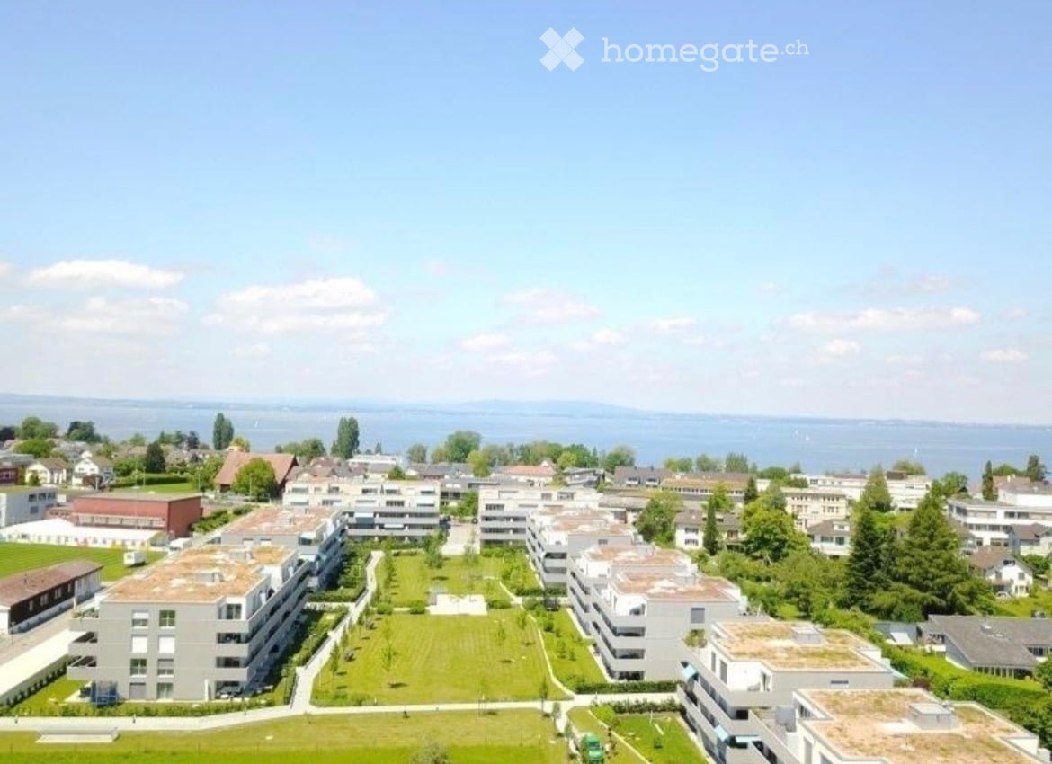 Aerial view of a residential complex with multiple apartment buildings, green spaces, and a body of water in the background.