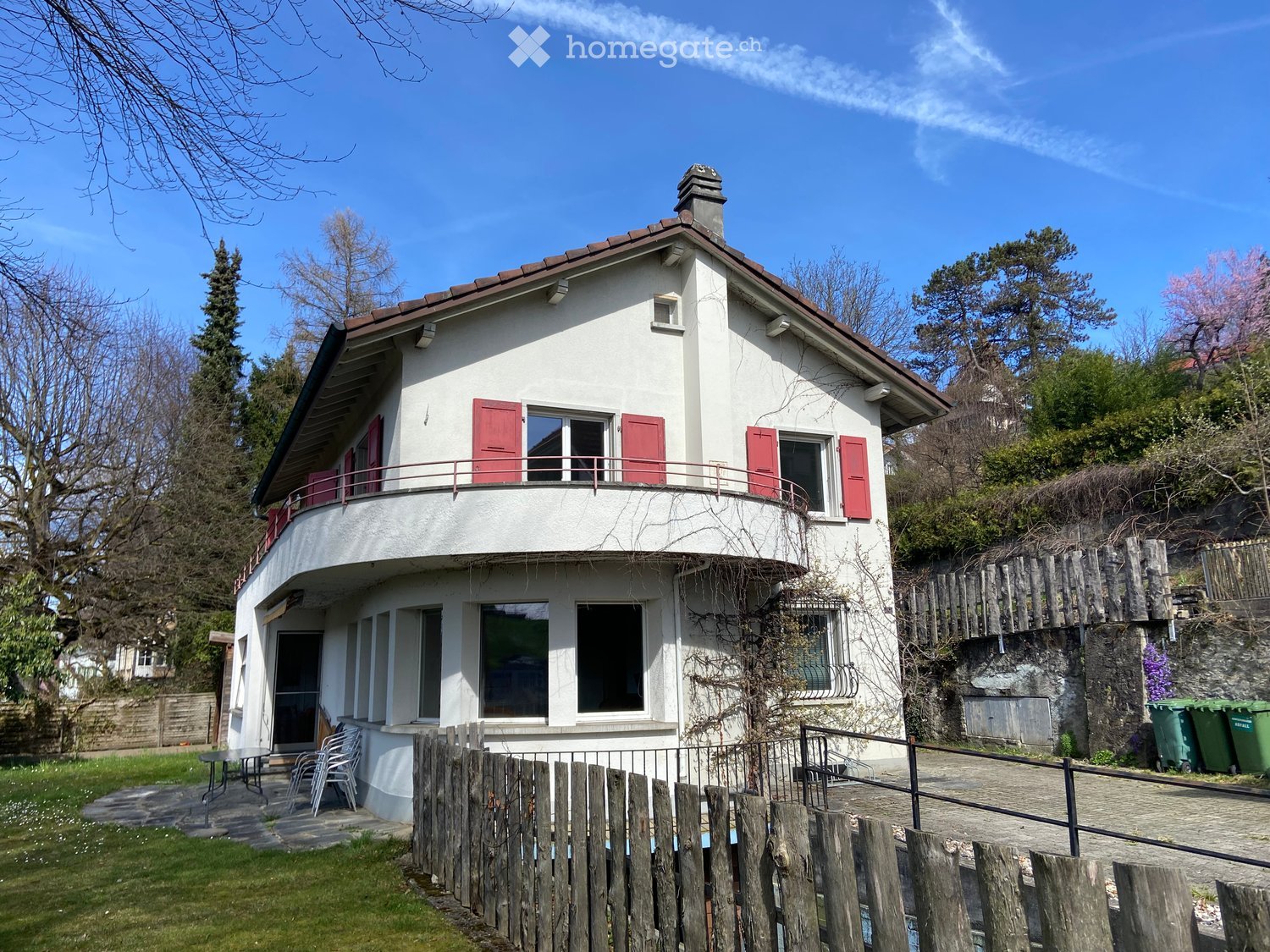Two-story house with white walls, red shutters, and a curved balcony on the upper level. The house features large windows and is surrounded by a wooden fence and greenery.