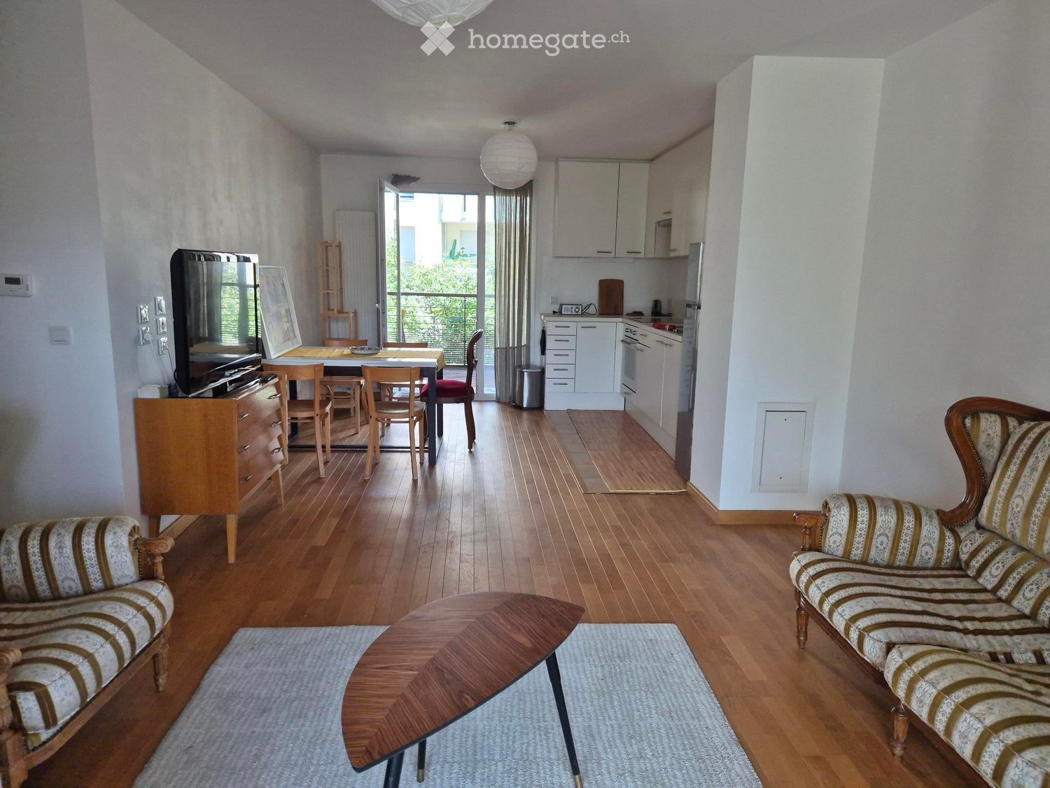 Open-plan living and dining area with wooden flooring, a sofa, coffee table, dining table with chairs, and a television. A small kitchen area is visible with cabinets, a refrigerator, and an oven. A balcony is accessible through large windows.