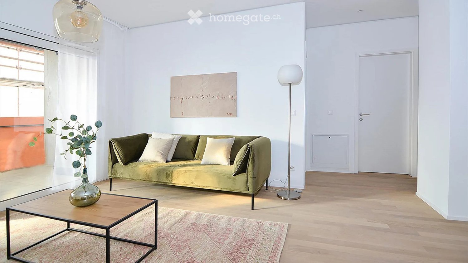 Modern living room with a green sofa, white throw pillows, a glass coffee table, a floor lamp, and a decorative vase with greenery on the coffee table. The room features light wooden flooring, white walls, and a large window with sheer curtains.