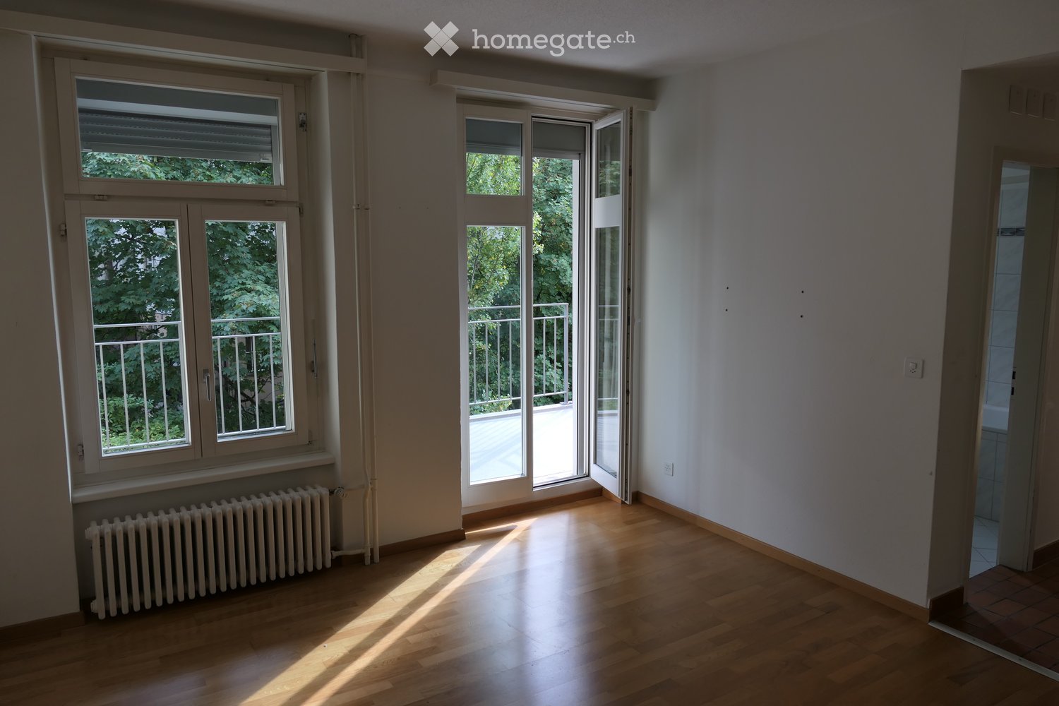 Empty room with wooden flooring, large windows, and a radiator. The room has a balcony accessible through a sliding glass door.