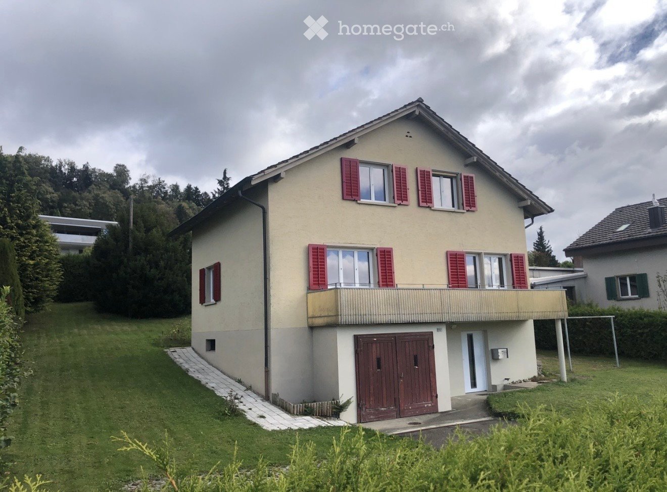 Two-story house with beige walls, red shutters, and a balcony on the upper floor. Features a garage entrance and a well-maintained lawn.