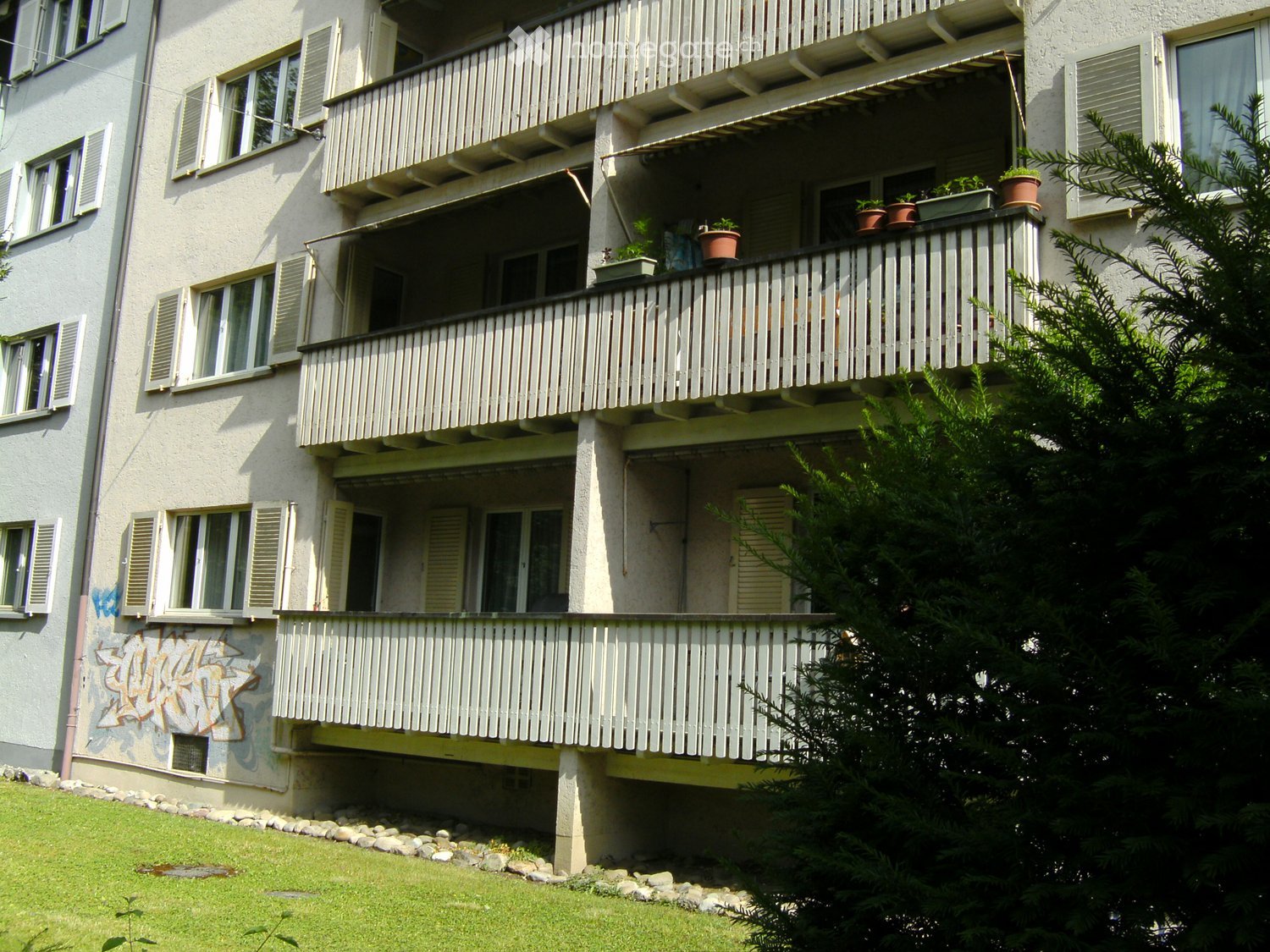 Multi-story residential building with multiple balconies, potted plants, and a grassy area in front.