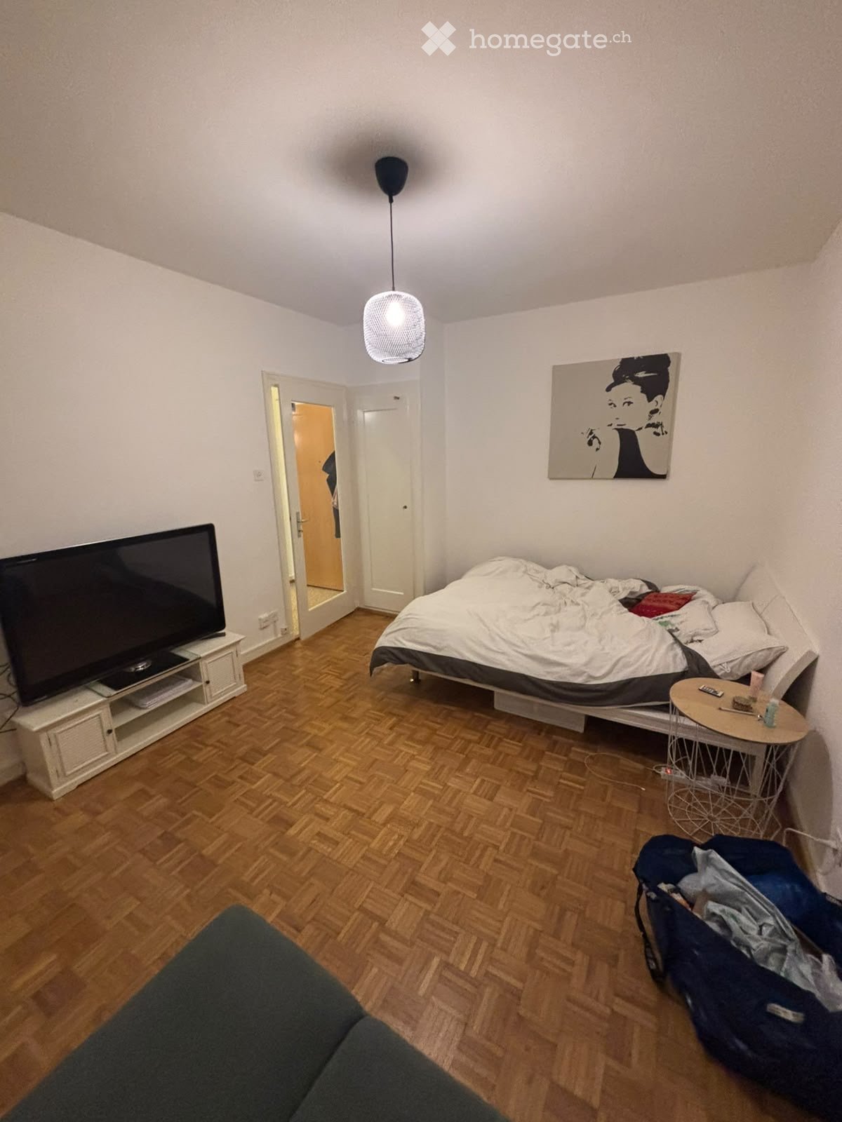 A modern bedroom with a wooden floor, a bed with white bedding, a nightstand, a small round table, a chair, a television on a stand, a hanging lamp, and a painting on the wall.