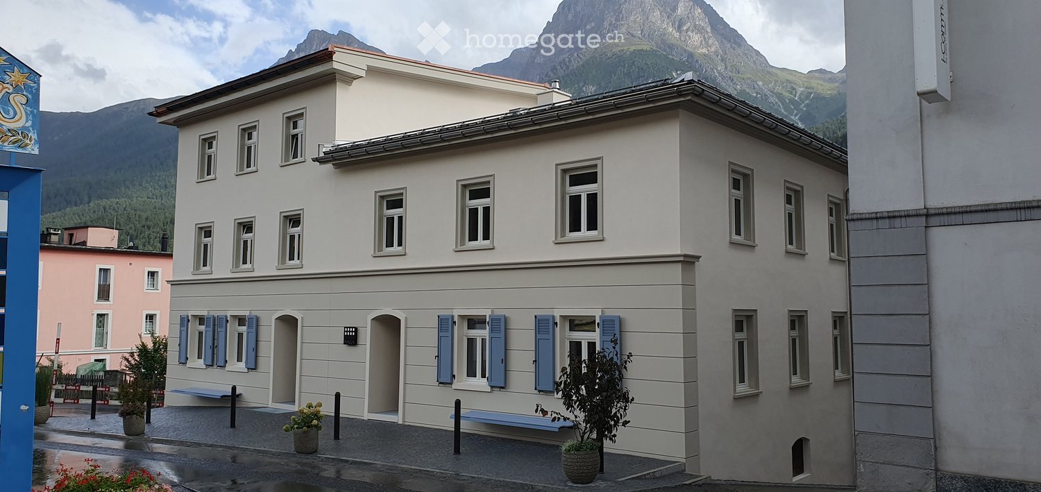 Two-story building with light gray facade, multiple windows, blue shutters, and a view of mountains in the background.