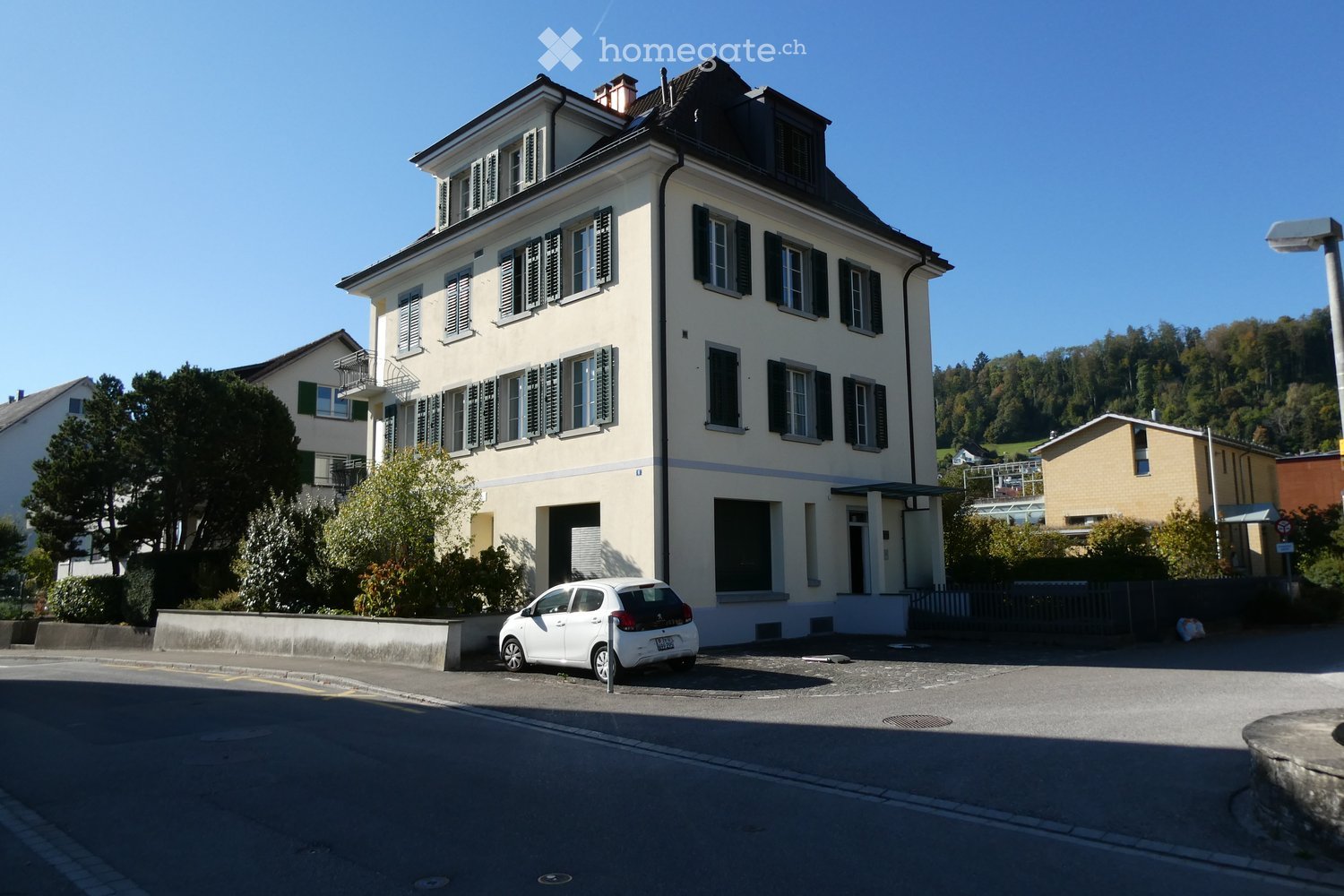 A three-story residential building with white walls, multiple windows with shutters, and a small balcony on the second floor. A white car is parked in front of the building.