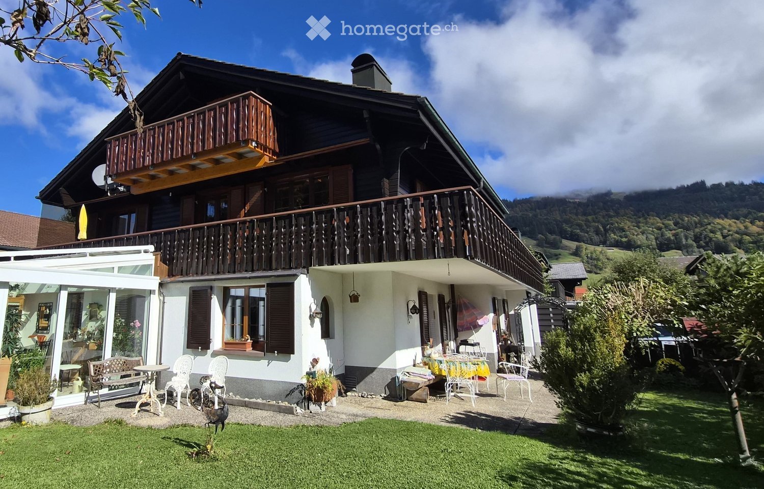 A two-story wooden house with balconies, surrounded by a well-maintained garden and outdoor seating area.