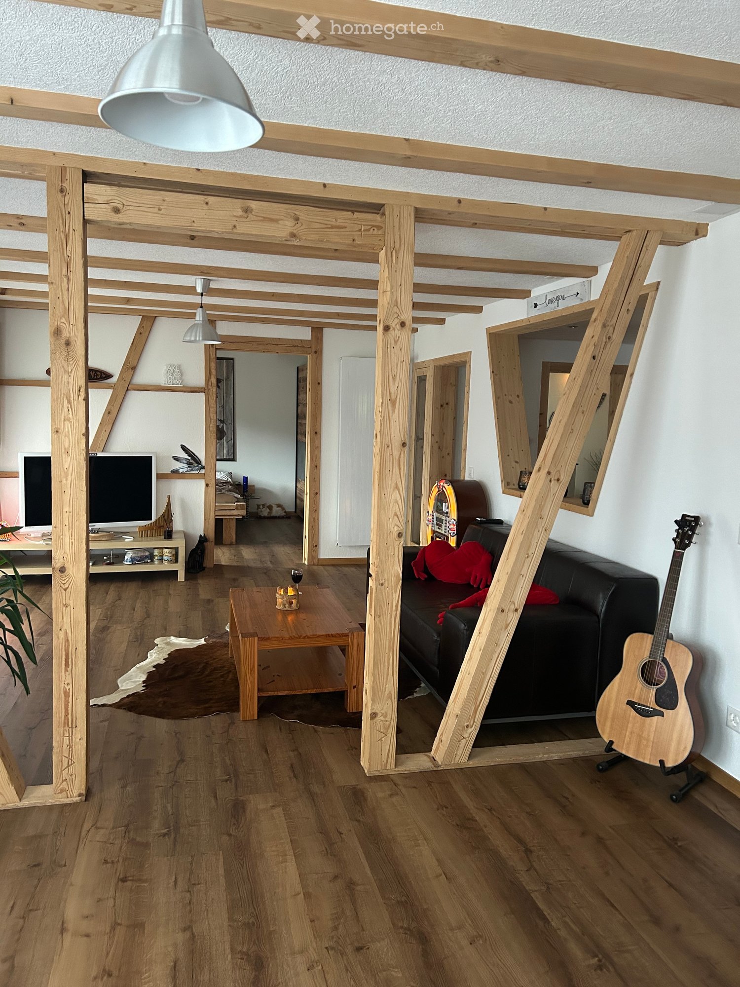 A modern living room with wooden beams, hardwood flooring, a black leather sofa, a wooden coffee table, a guitar, and a television on a stand.