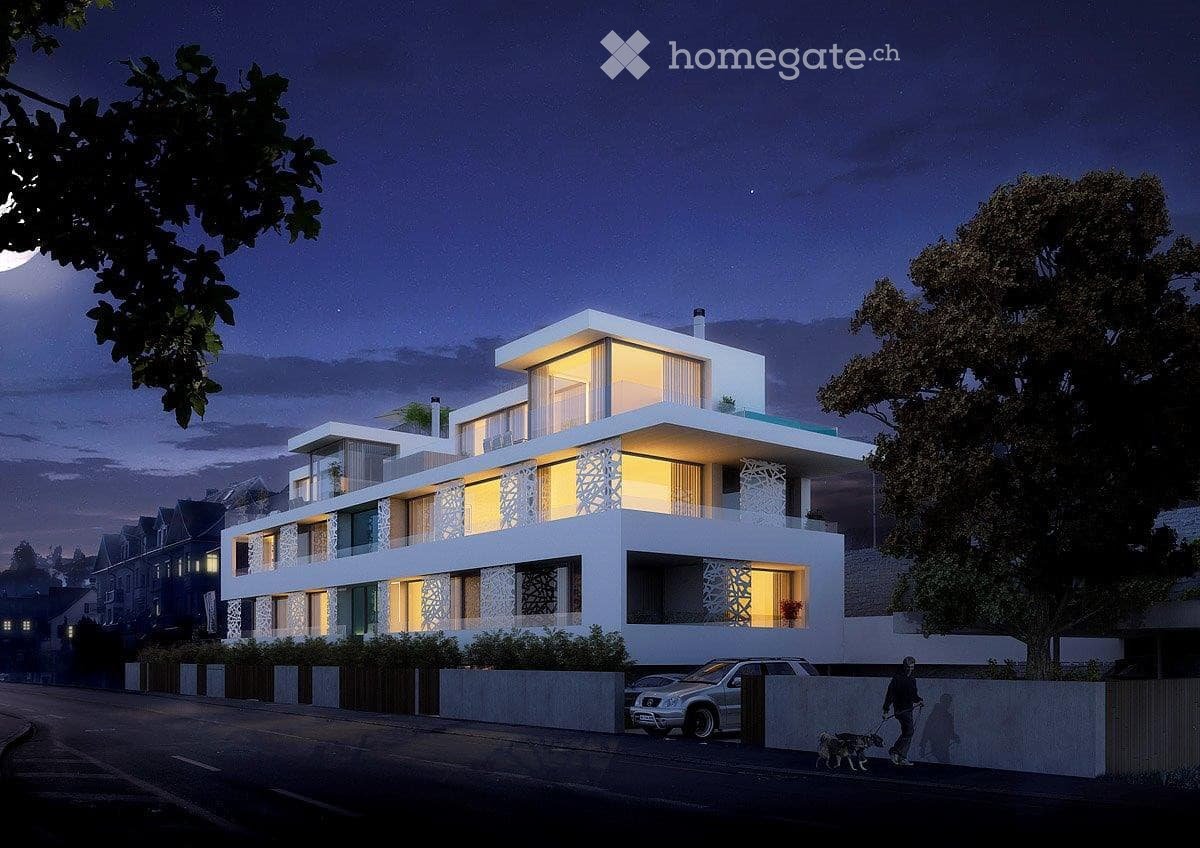 Modern, multi-story residential building with white exterior, large windows, and illuminated interiors. Features include balconies, a raised ground floor, and a parking area with a car visible.