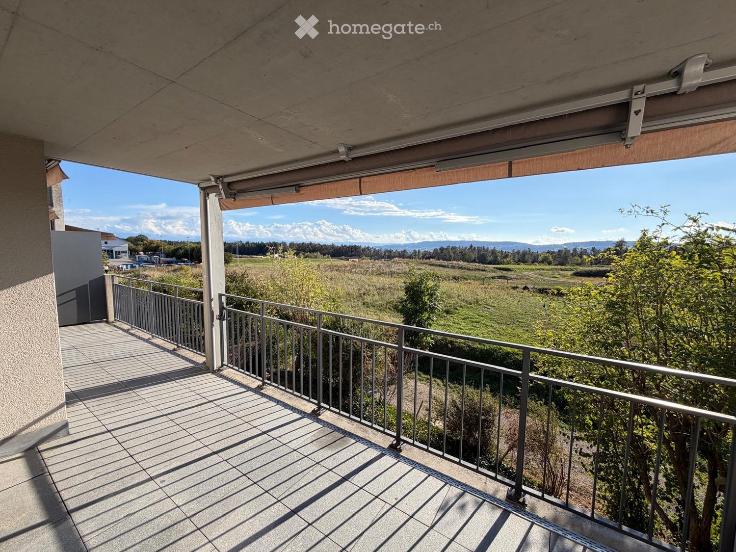 Balcony with tiled floor, metal railing, and a retractable awning, overlooking a green field and distant mountains.