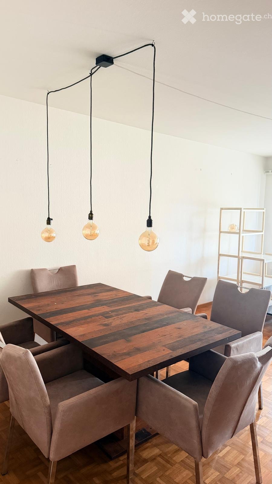 Dining area with a wooden table and six upholstered chairs, pendant lights hanging from the ceiling, and a modern shelving unit in the background.