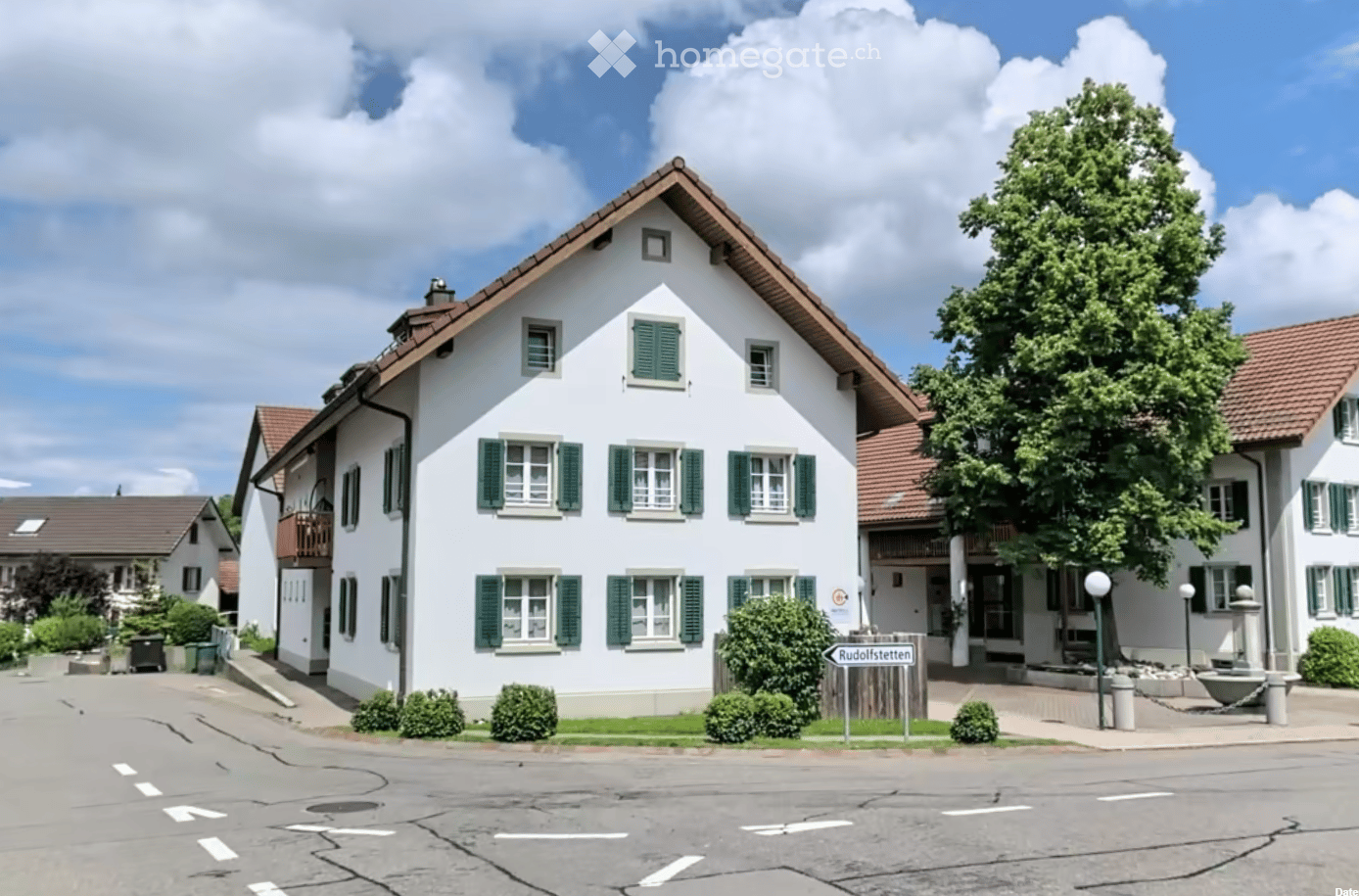 A two-story white house with green shutters and a brown roof, surrounded by greenery and a street sign reading 'Rudolfstetten'.