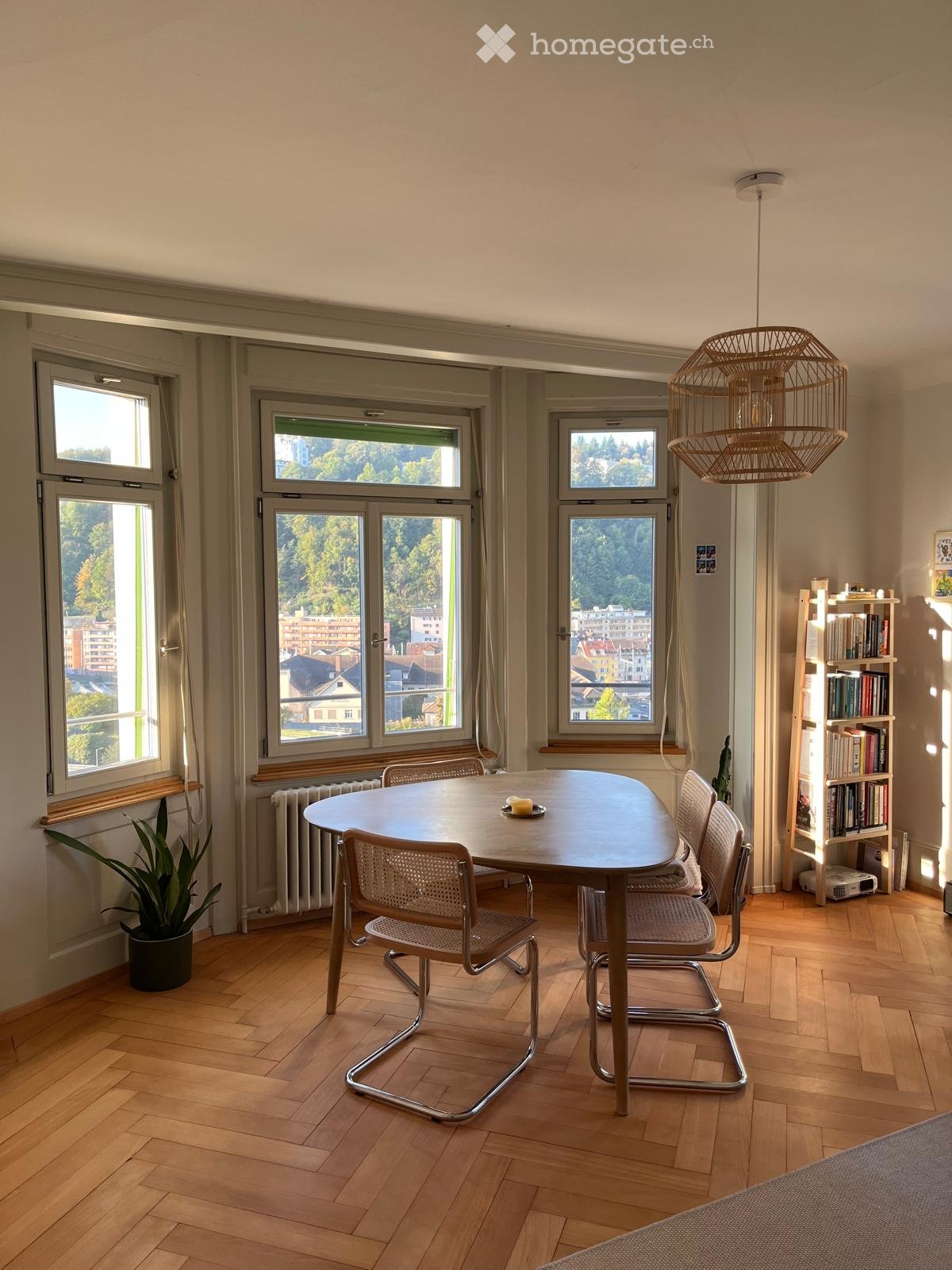 Bright dining area with wooden floor, large windows overlooking a cityscape, round wooden table with four chairs, bookshelf, potted plant, and a hanging woven light fixture.