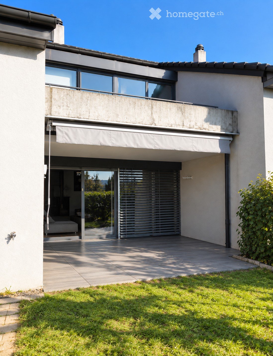 Modern two-story house with large windows, a balcony, and a covered entrance area. The house features a clean, contemporary design with a raised ground floor and a well-maintained lawn in the foreground.