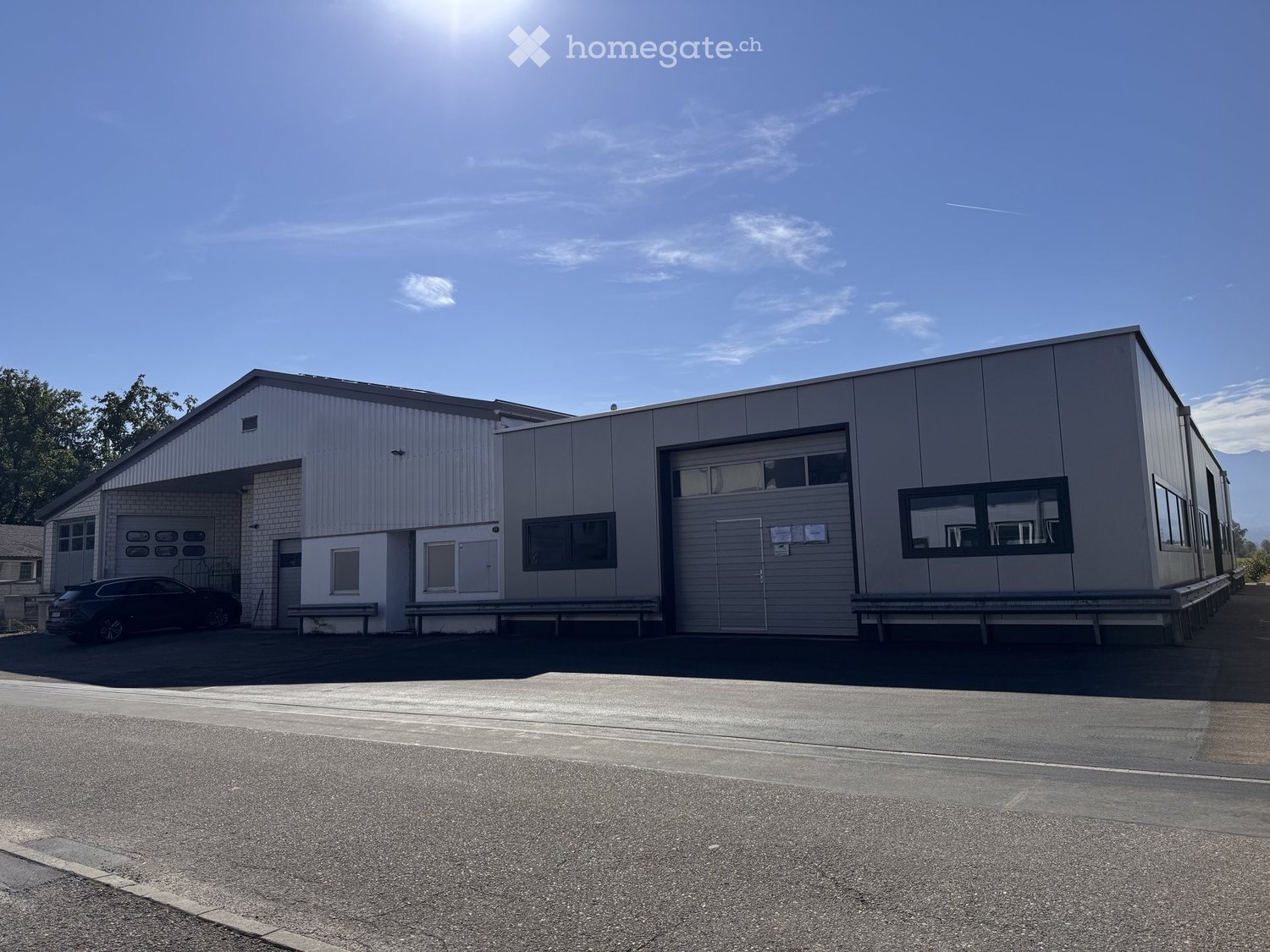 Modern industrial-style building with a large garage door, multiple windows, and a flat roof, situated under a clear blue sky.