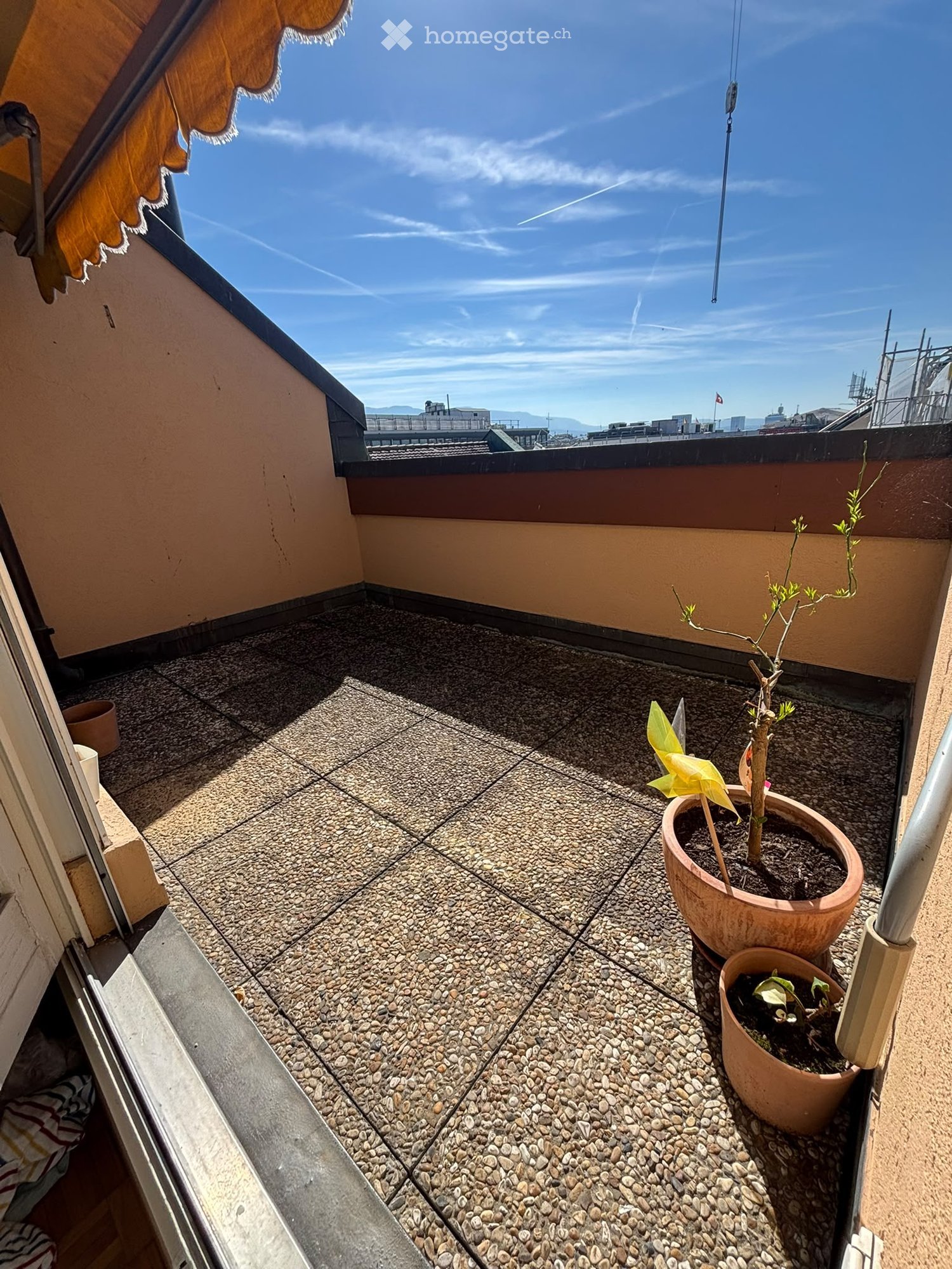 Small balcony with a gravel floor, two potted plants, and a view of the city and mountains under a clear blue sky.