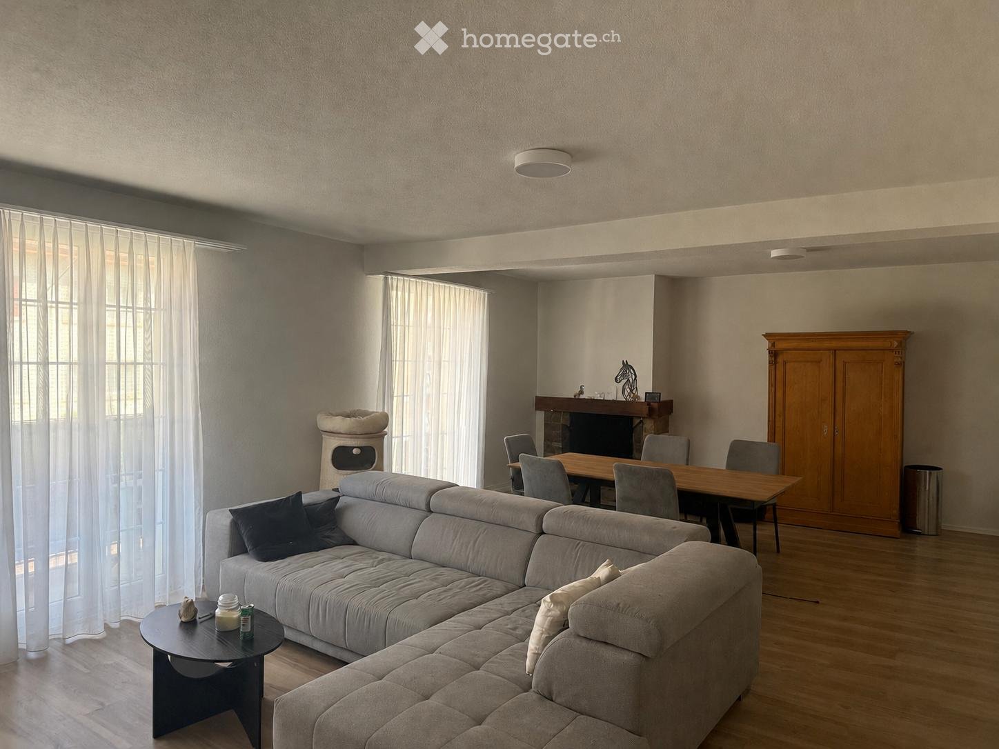 Modern living and dining area with a grey sectional sofa, coffee table, dining table with chairs, and a wooden cabinet. The space features large windows with sheer curtains and a fireplace.