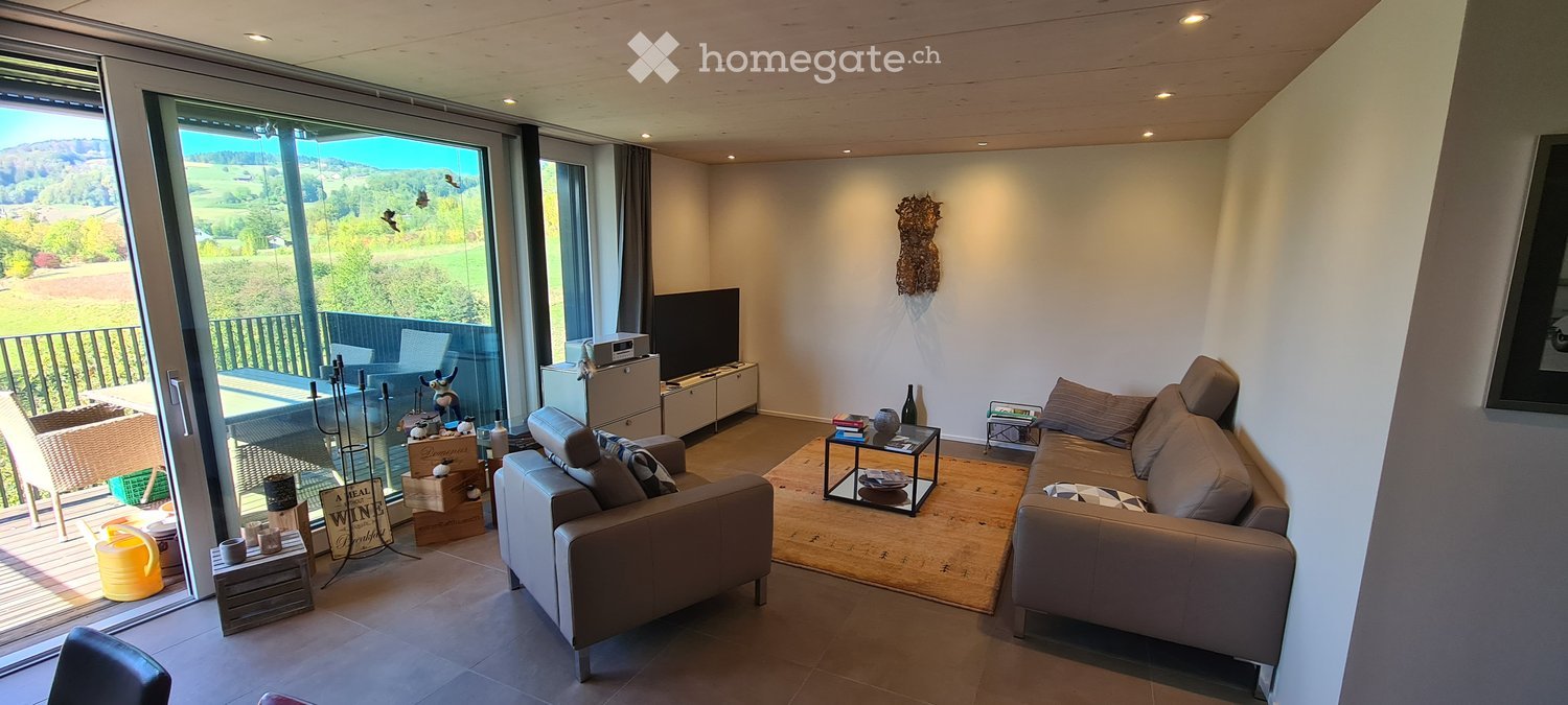 Modern living room with large windows offering a scenic view, featuring a grey sectional sofa, coffee table, side table, television, and a small table with decorative items.
