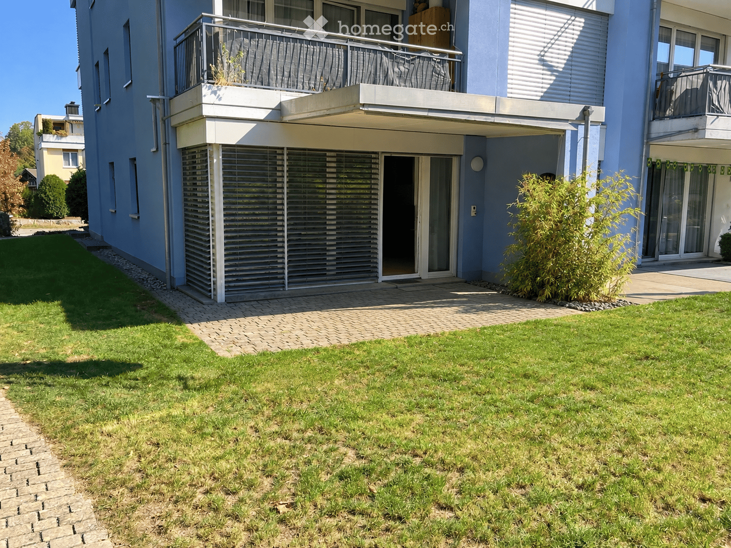 Well-maintained garden with a paved pathway leading to a modern building entrance, featuring balconies and large windows.