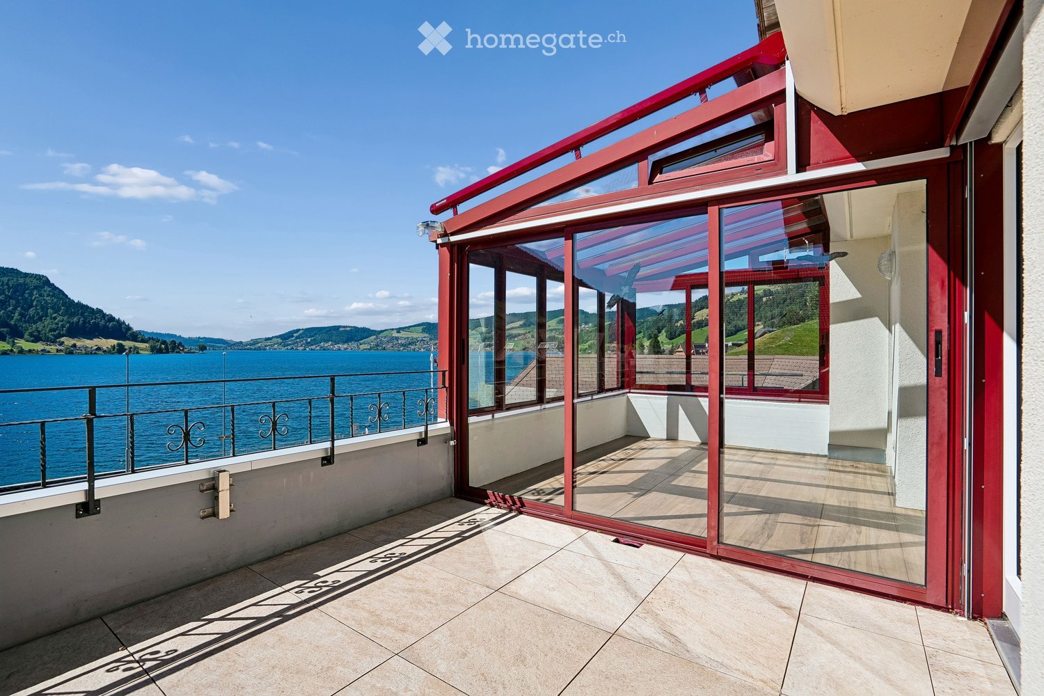 Balcony with large glass sliding doors, red frame, tiled floor, metal railing, and scenic lake view.
