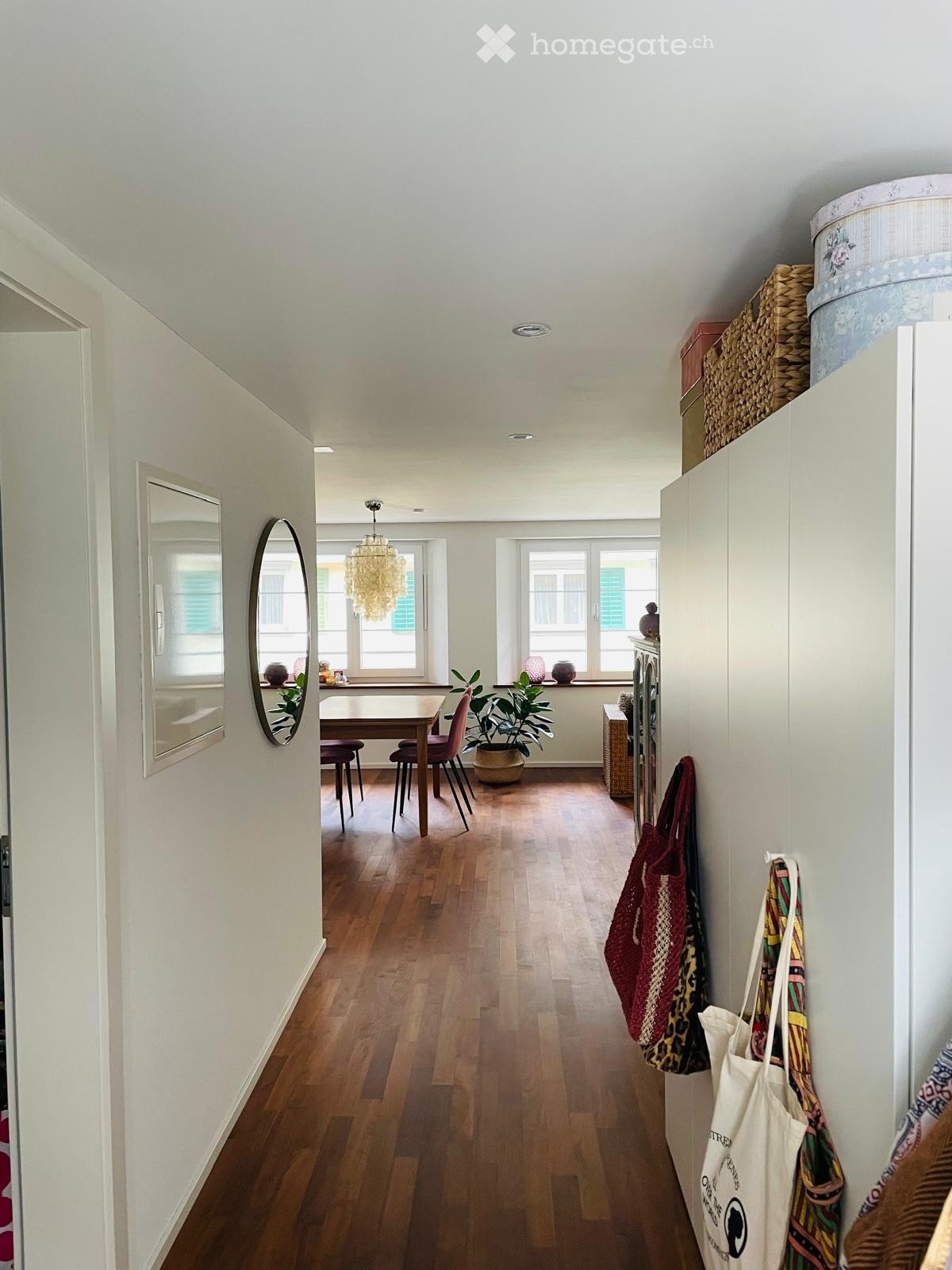 Bright hallway with wooden flooring, white walls, and large windows at the end. A dining area is visible through an open doorway, featuring a table, chairs, and a chandelier hanging from the ceiling.