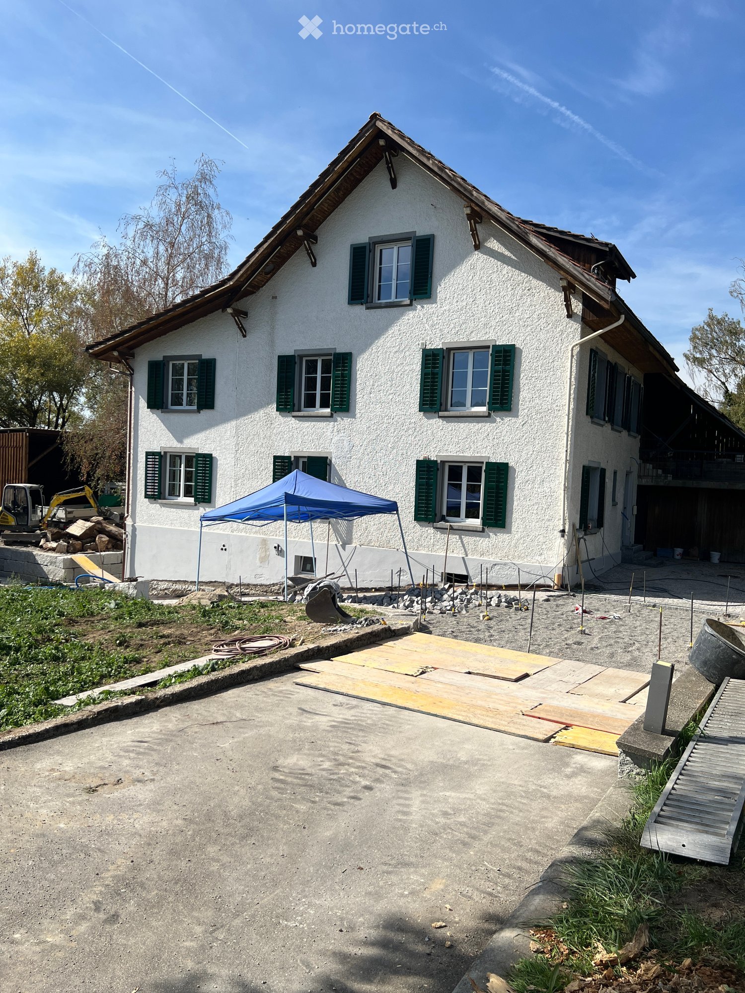 Two-story white house with green shutters and multiple windows, under construction with a blue canopy and construction materials nearby.