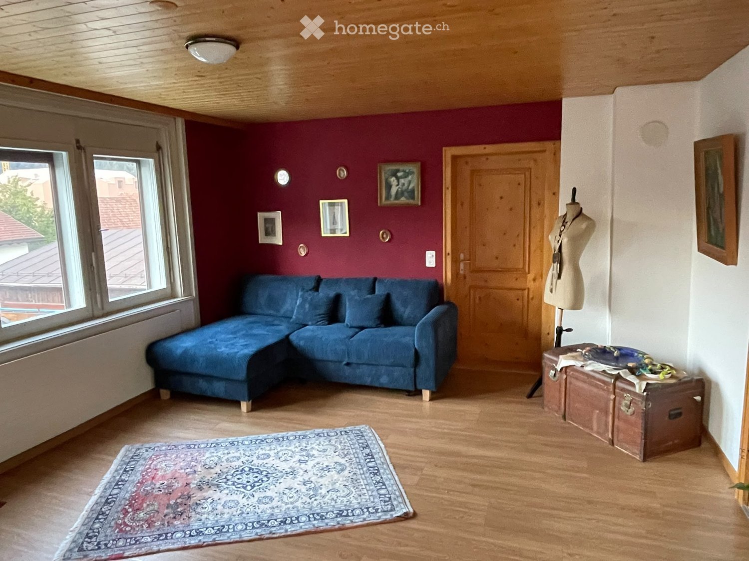 Living room with wooden floors, a blue sectional sofa, a patterned rug, several picture frames on the wall, and a mannequin with accessories on a suitcase.