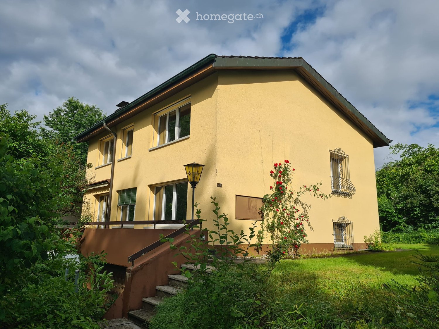 Two-story yellow house with multiple windows, a staircase leading to the entrance, and surrounding greenery.