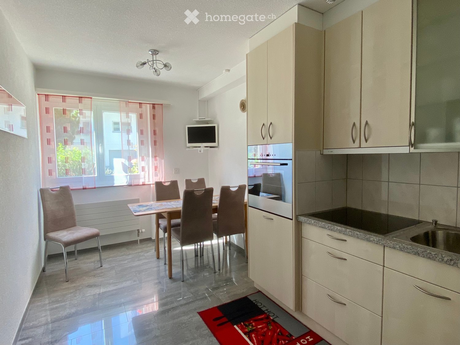 A modern, well-lit kitchen and dining area with a dining table, six chairs, kitchen cabinets, an oven, a sink, and a television mounted on the wall. The space features a tiled floor and a red and black runner.