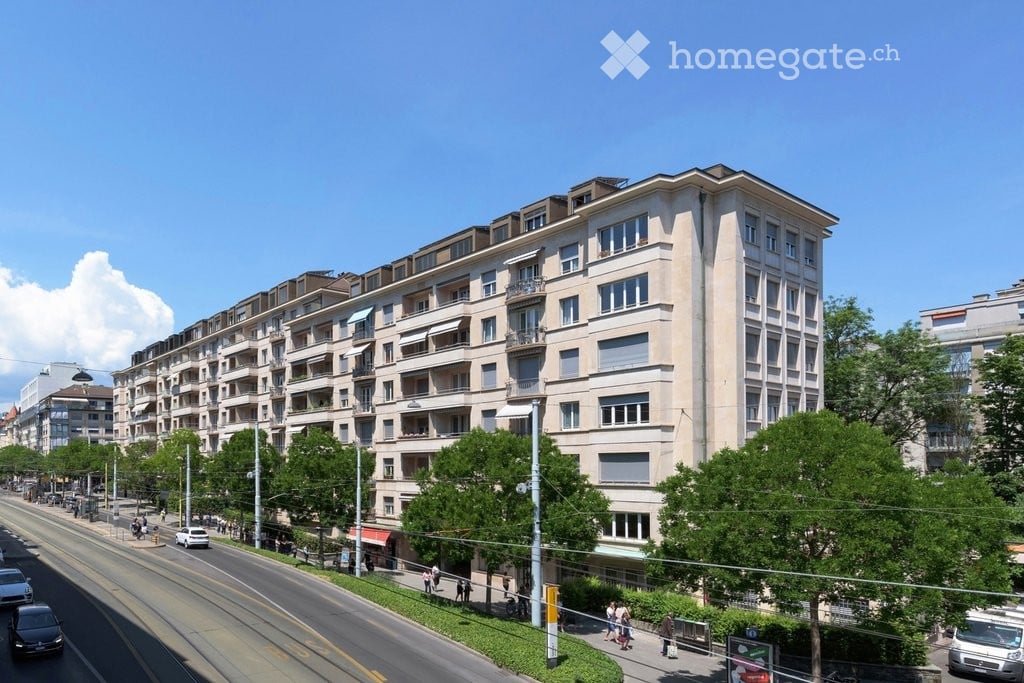Multi-story residential building with balconies and large windows, located on a busy street with trees and pedestrians.